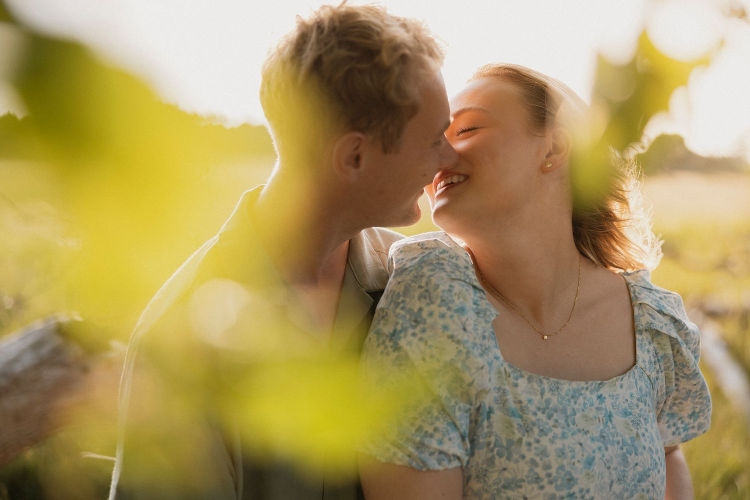 Young couple sharing a romantic moment kissing in a sunlit field surrounded by lush green foliage.