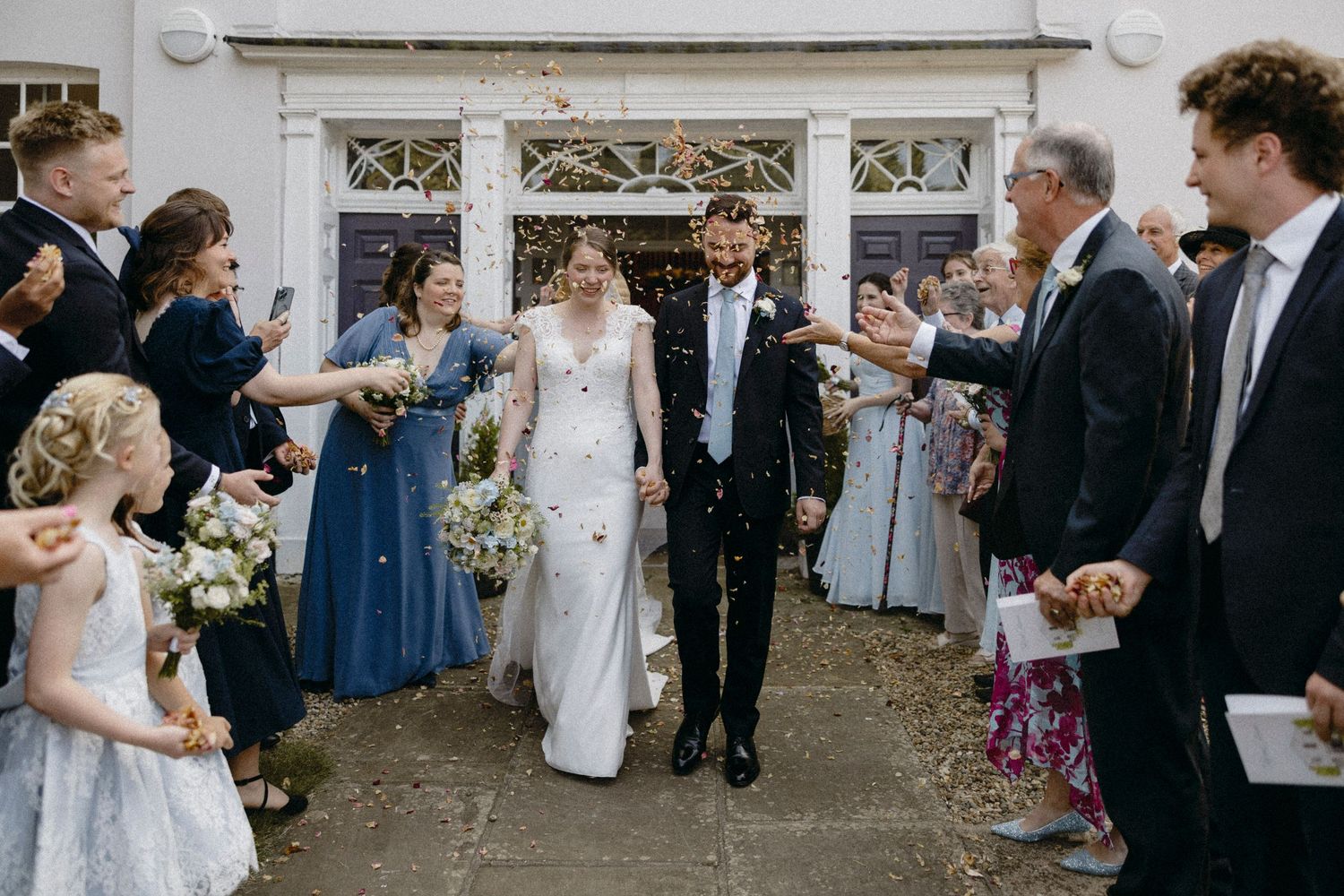 Bride and groom walk through confetti shower surrounded by smiling guests outside a white wedding venue.