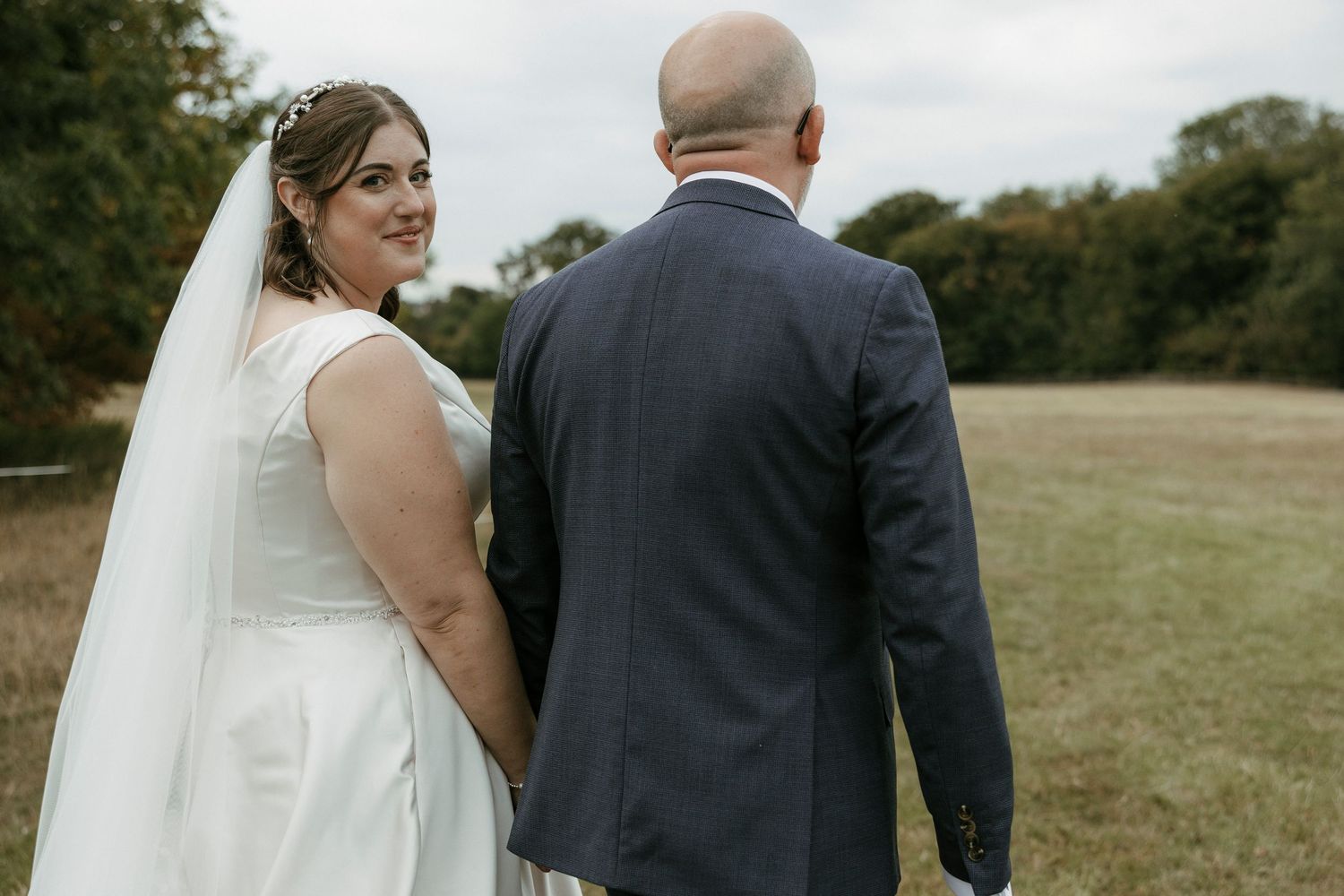Bride in white gown and veil looks back smiling while holding grooms hand in scenic field.