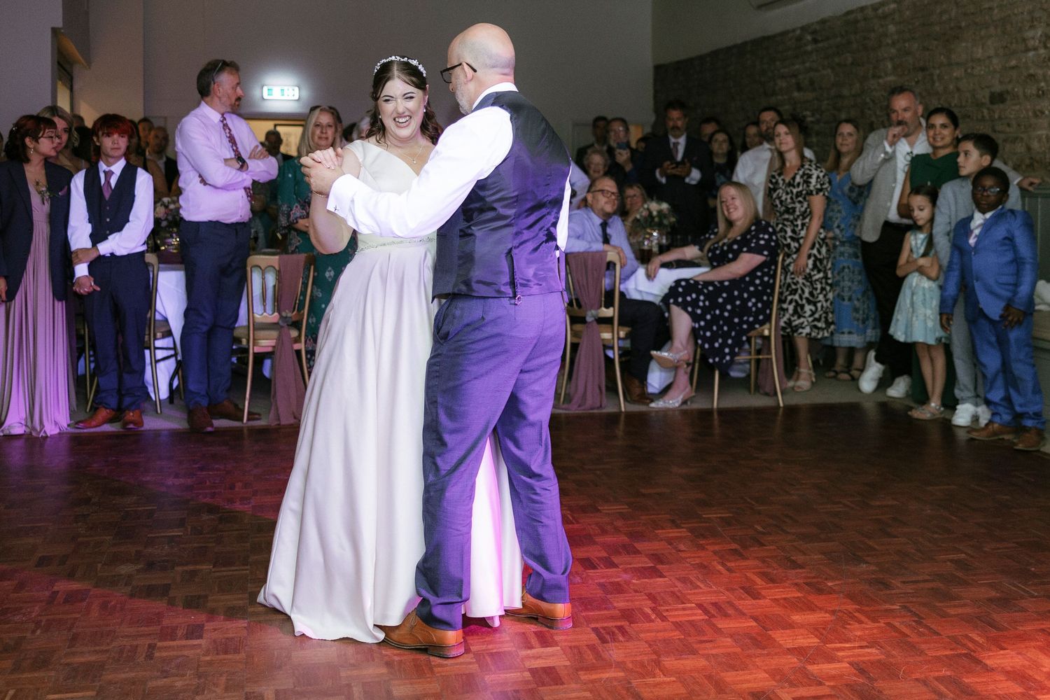 Bride and groom share their first dance at wedding reception, surrounded by guests watching on a wooden dance floor.