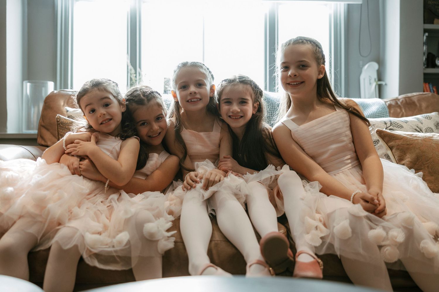 Five young flower girls in pink tulle dresses smiling together on a sofa before a wedding.
