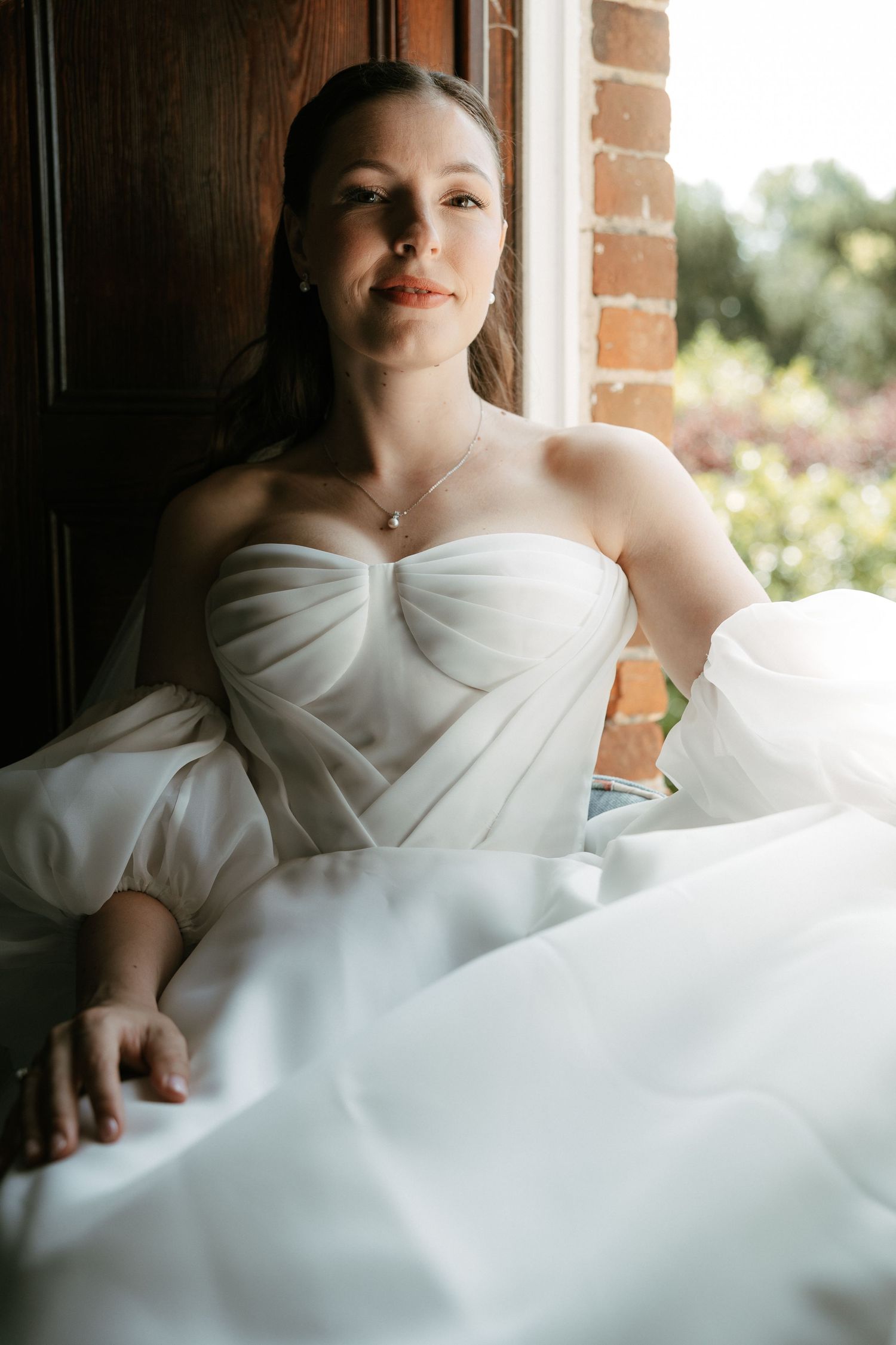 Bride in a white strapless tulle wedding gown seated by a window, wearing a delicate necklace, with soft natural light.