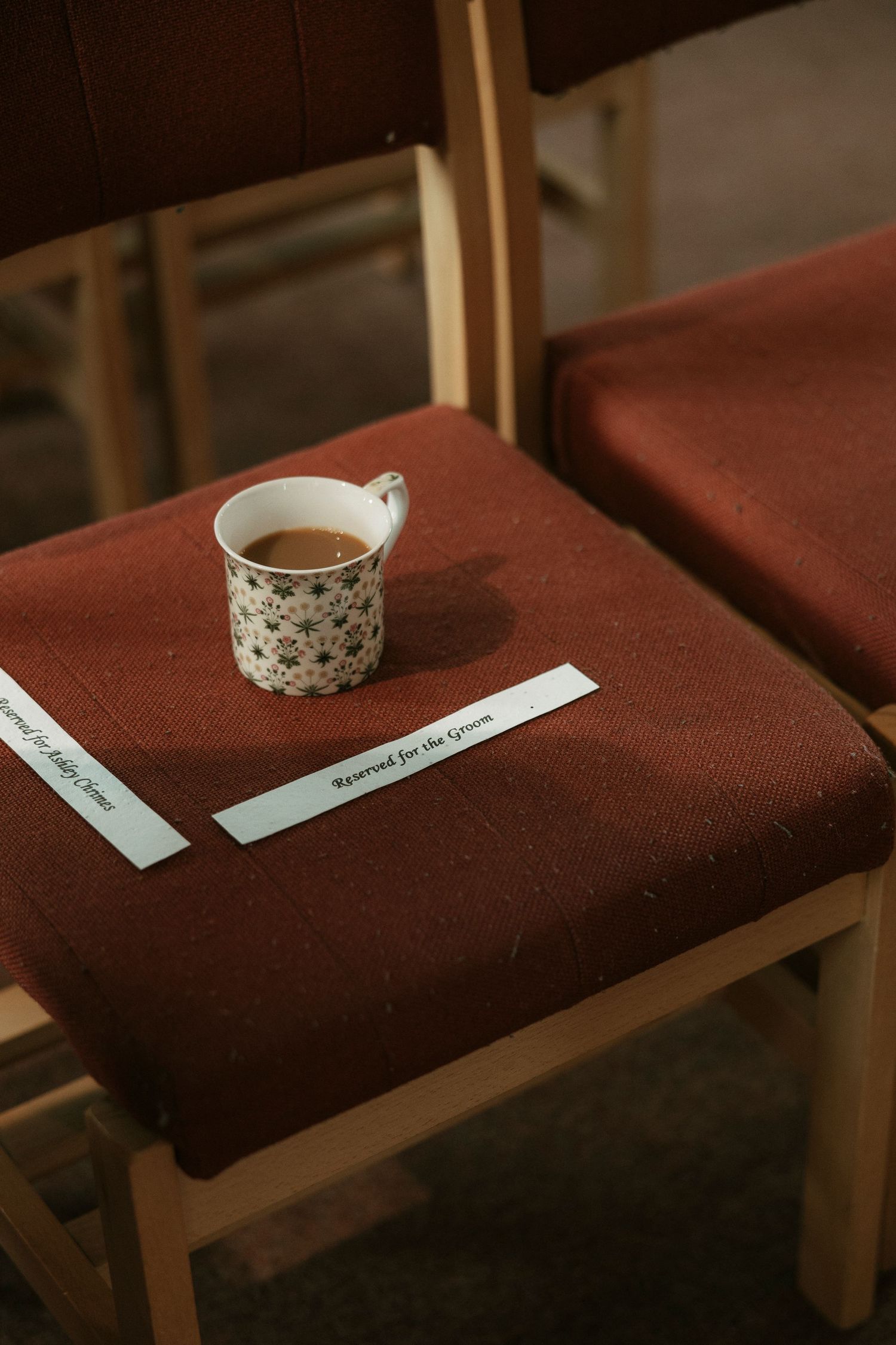 A floral ceramic mug of coffee sits on a reserved red fabric chair at a wedding ceremony venue.