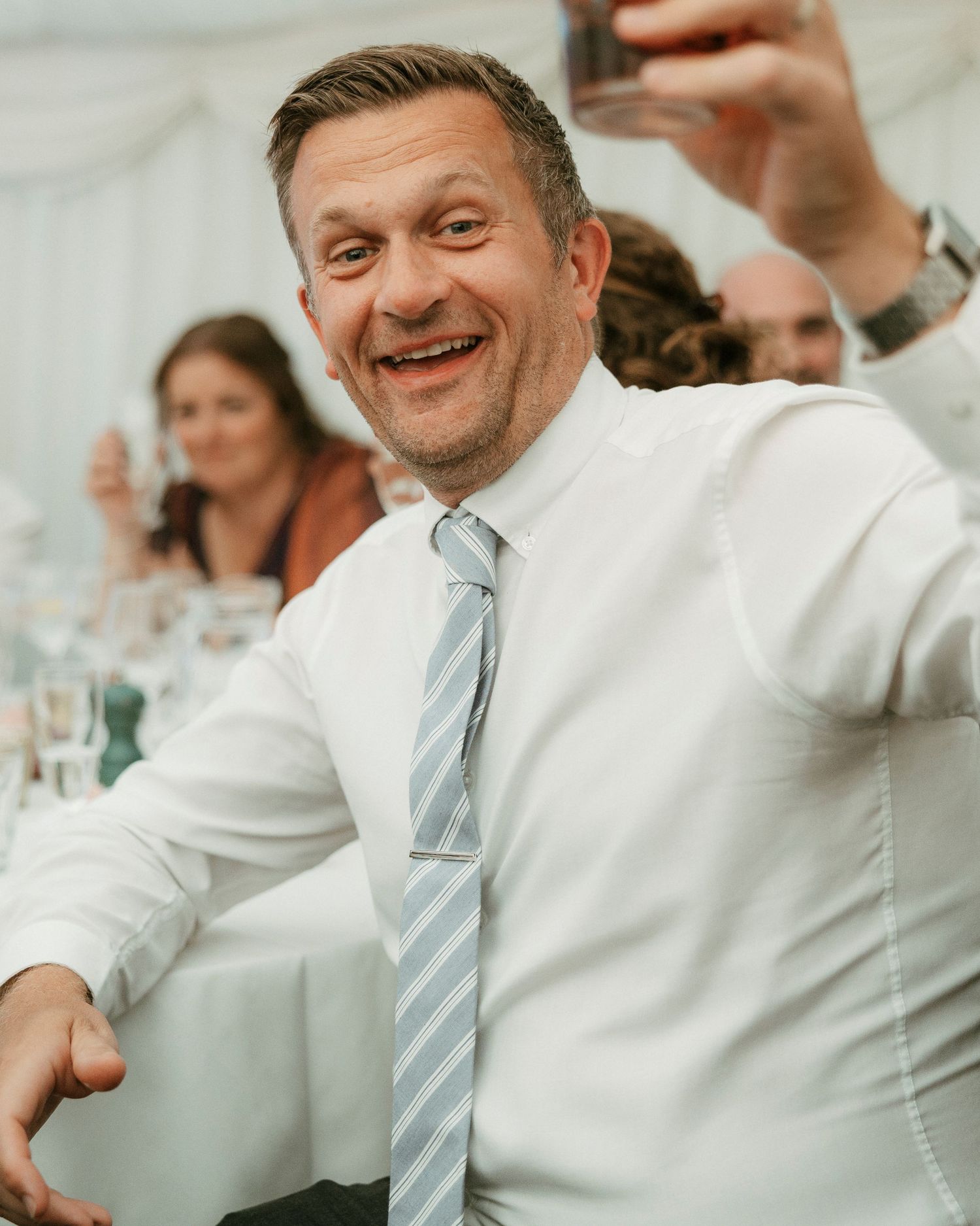 Smiling man in white shirt and striped tie raising a glass in a toast at a wedding reception.