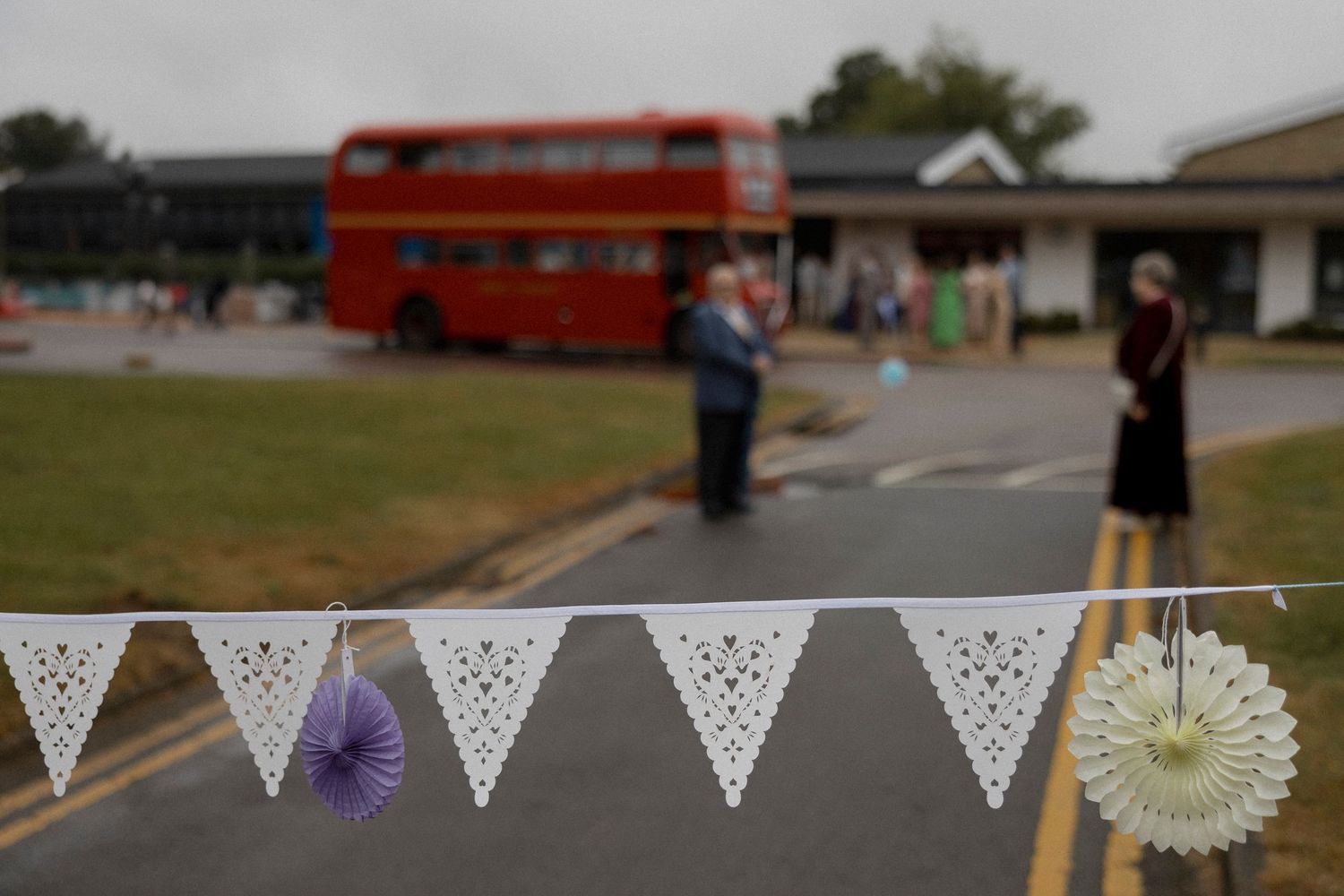 White lace bunting foreground with red double-decker bus and guests at outdoor garden party event.