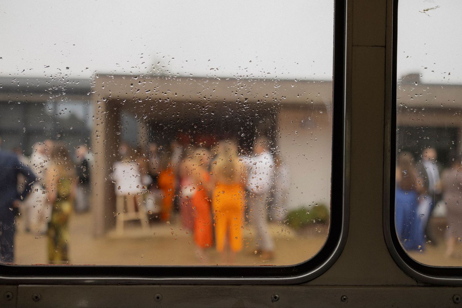 Rain-streaked window view of blurred figures in orange prison jumpsuits gathered outside a correctional facility.