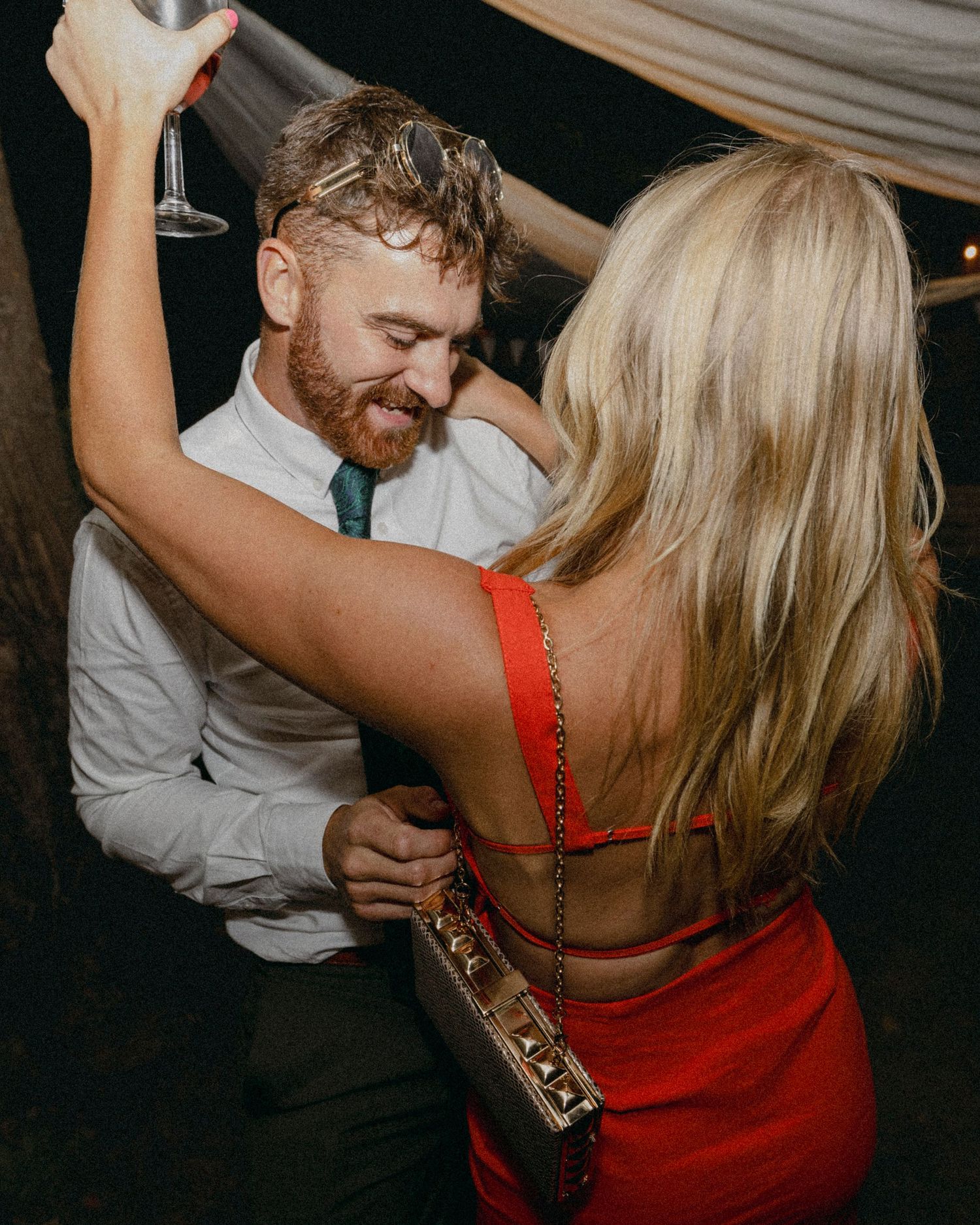 Couple dancing closely at a wedding reception, man in white shirt with green tie, woman in red backless dress.