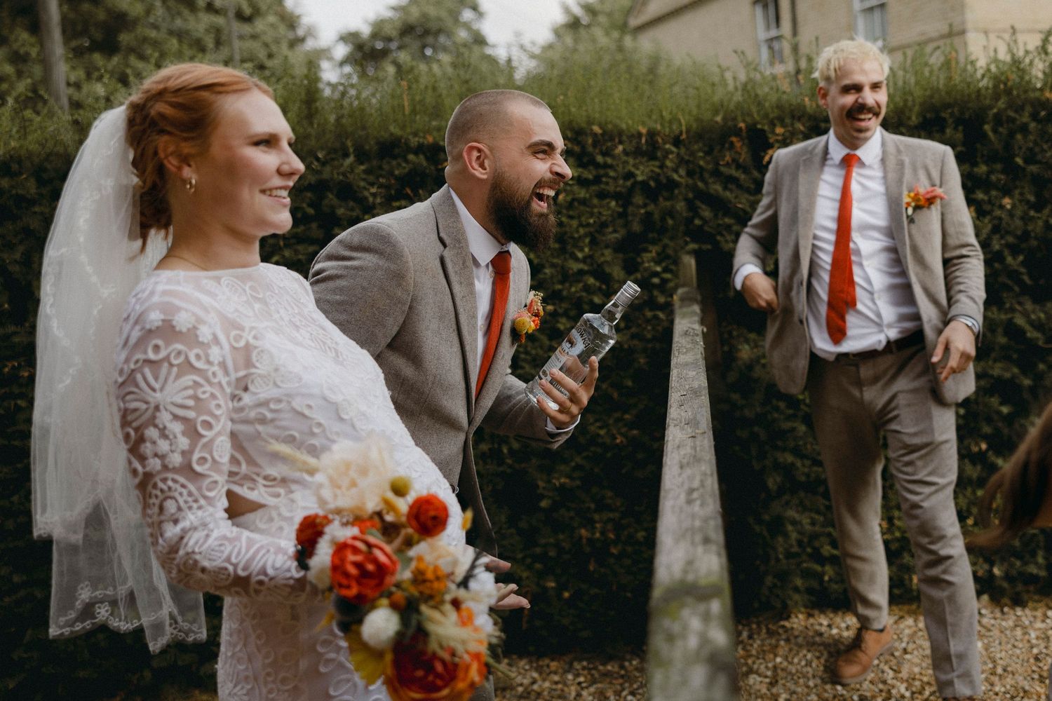 Bride in lace dress holding bouquet laughs with groom in gray suit as guest photobombs at outdoor wedding.