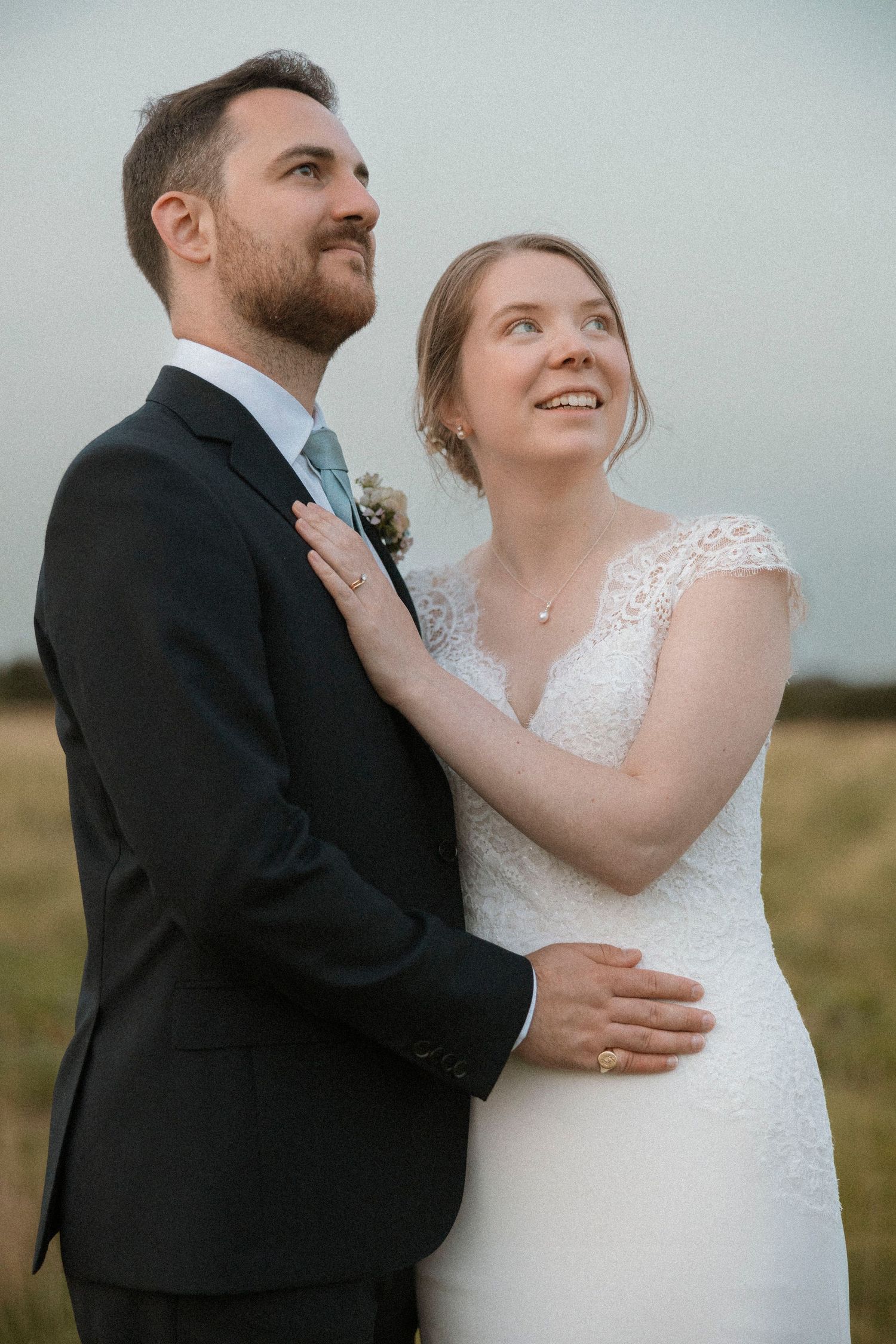 A bride and groom in wedding attire embrace outdoors in a field, both gazing upward with joyful expressions.