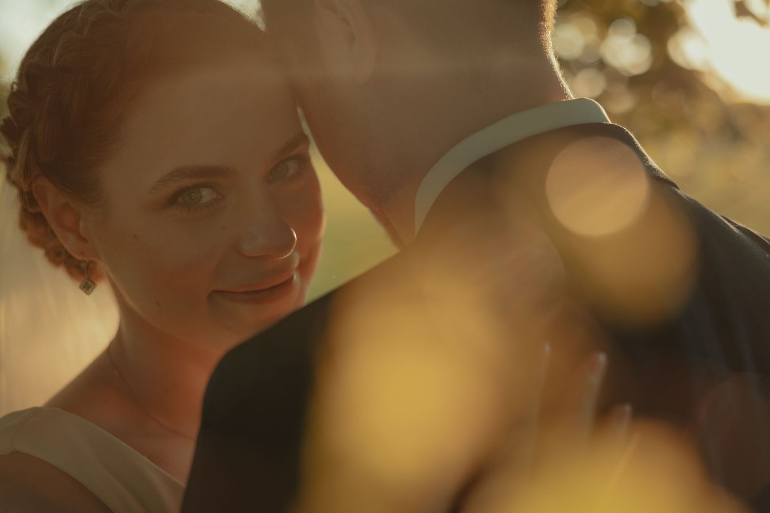 A bride smiles softly in golden sunset light, leaning close to her groom in a romantic outdoor wedding portrait.