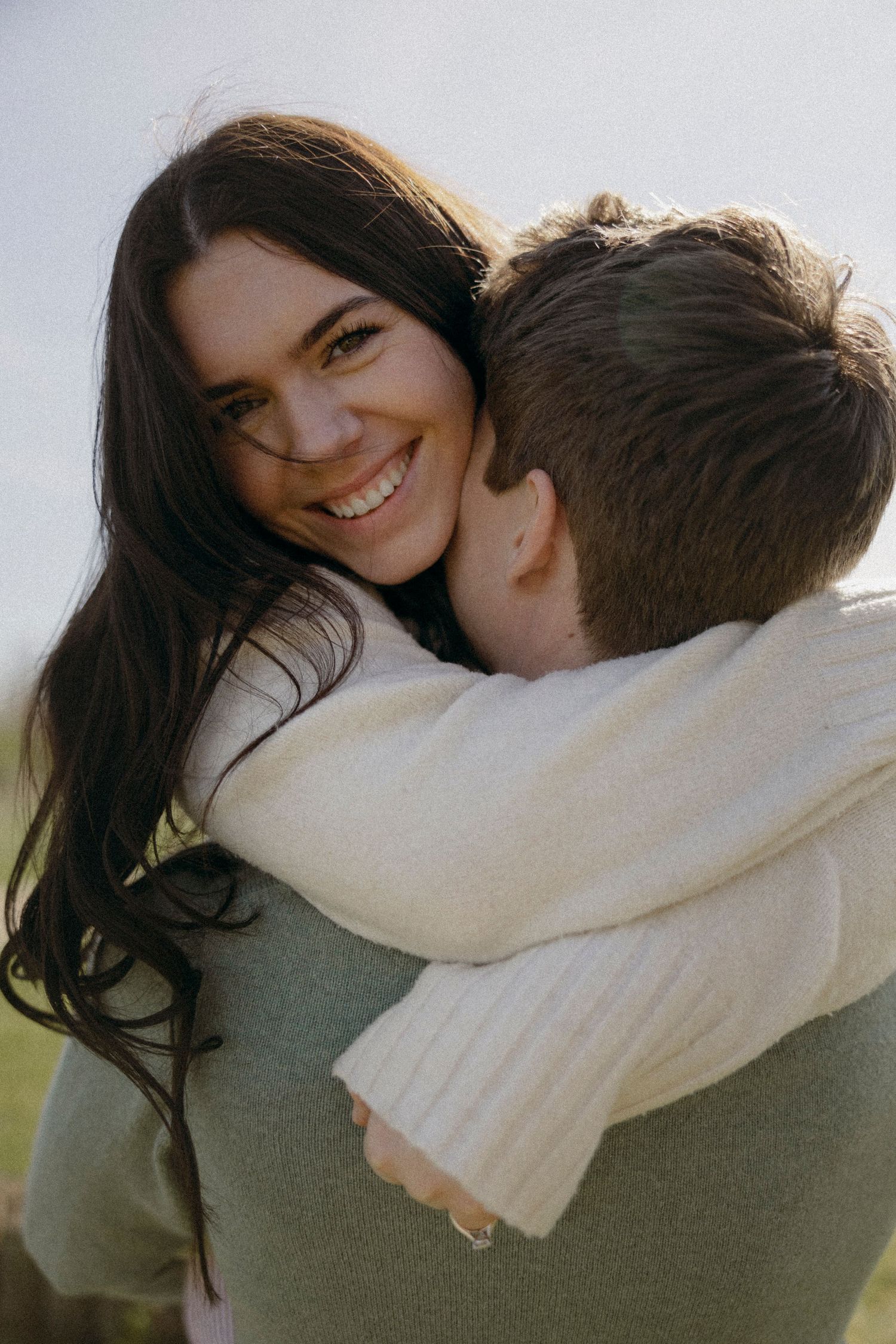 A smiling brunette woman hugs a man in a cream sweater outdoors in a warm, candid couple embrace.
