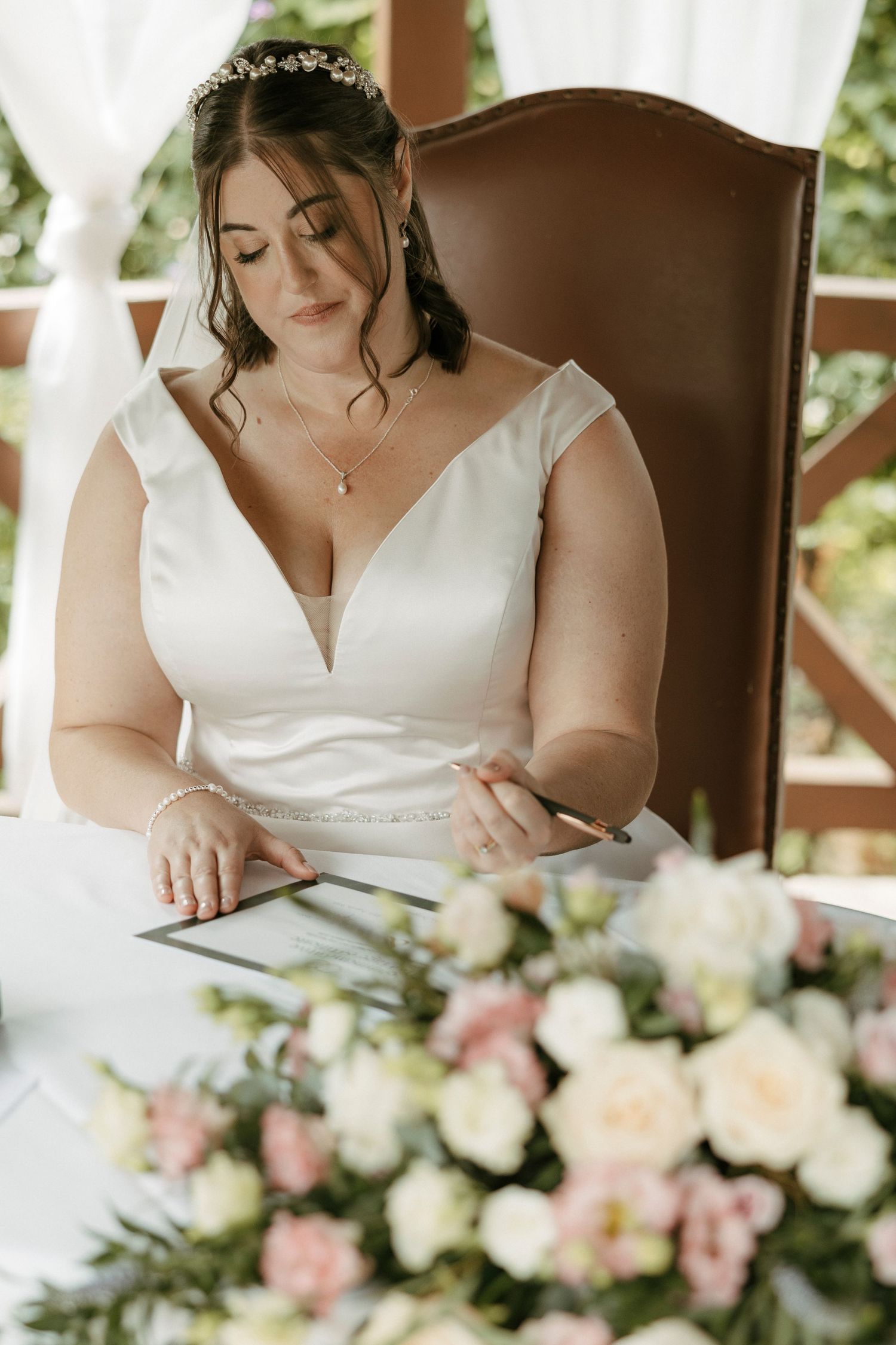 Bride in off-shoulder satin gown signing wedding register at decorated table with floral arrangement.