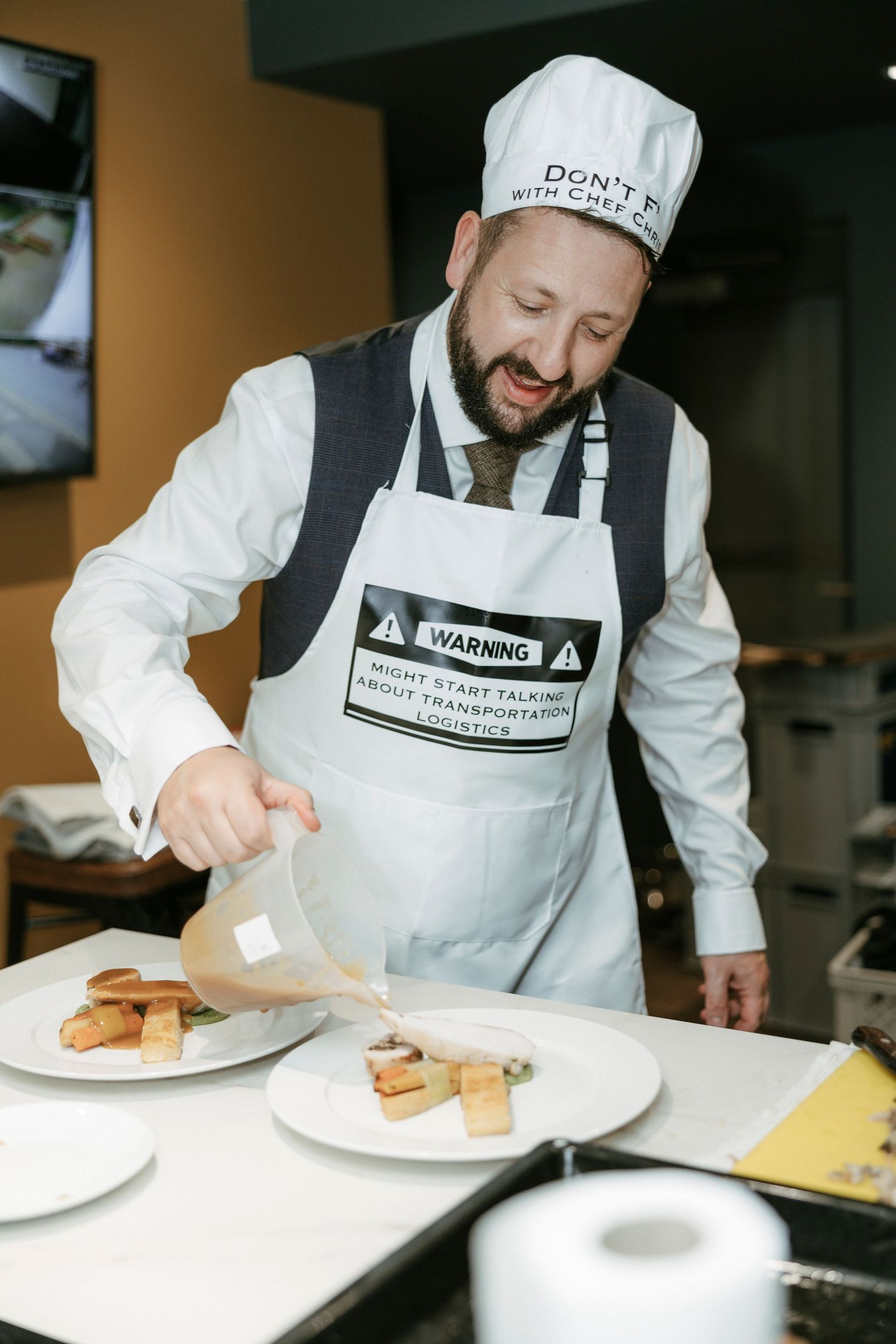 A smiling chef in a humorous apron and hat plates gourmet dishes in a professional kitchen setting.