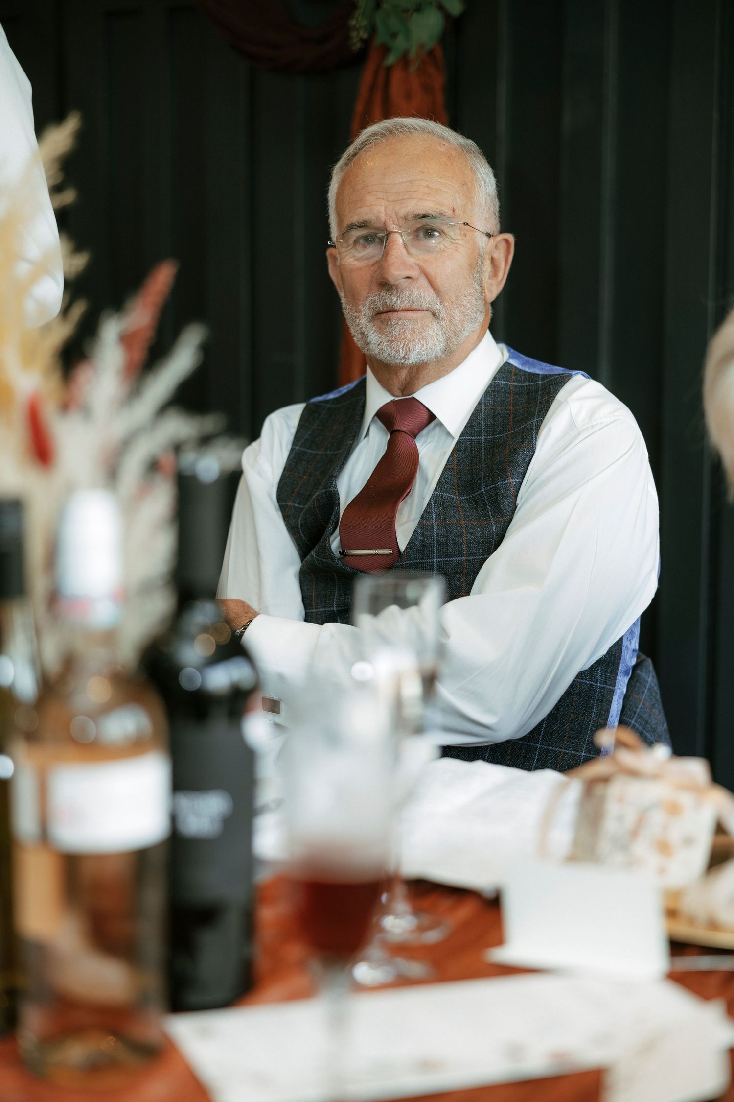 Elderly man with glasses and beard wearing vest and red tie sits at a restaurant table with wine glasses.
