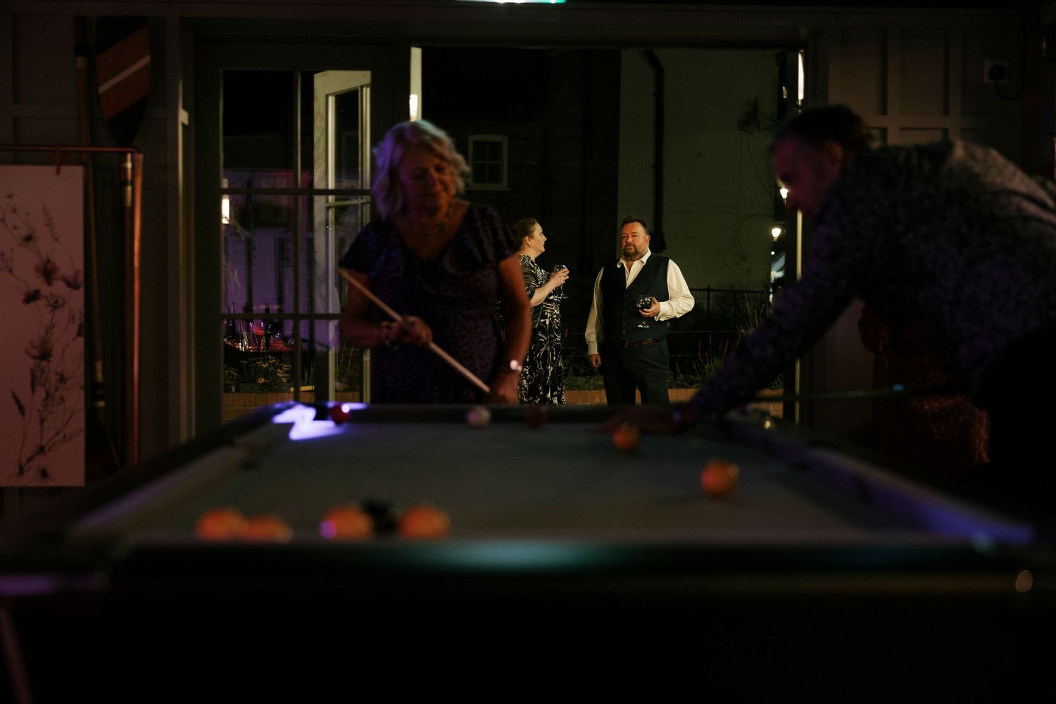 People playing pool billiards in a dimly lit room at a social gathering around a glowing table.