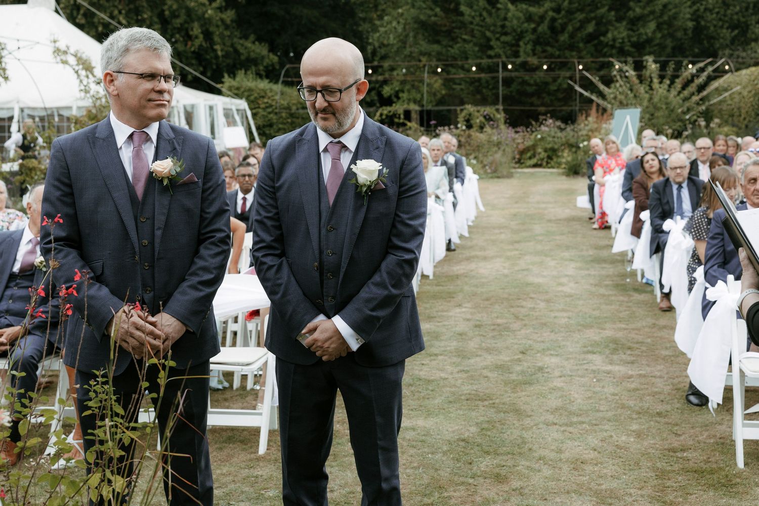 Two grooms in navy suits stand at the altar during an outdoor wedding ceremony on a lush green lawn.