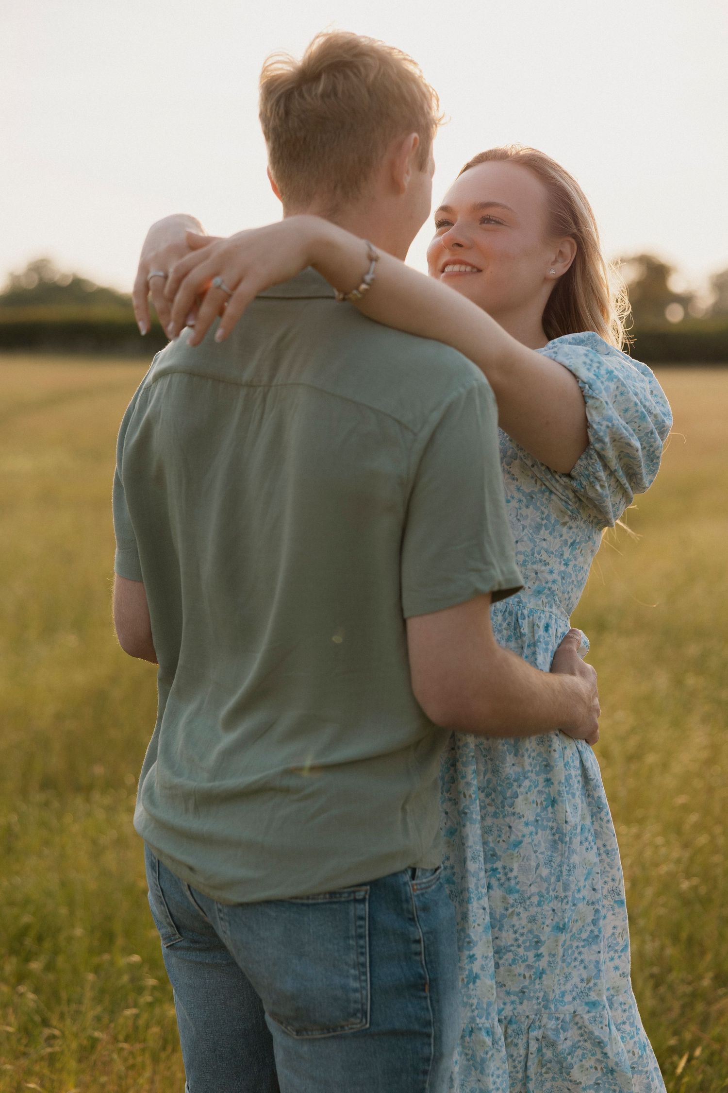 Couple embracing in a golden wheat field at sunset, woman in floral dress smiling warmly.