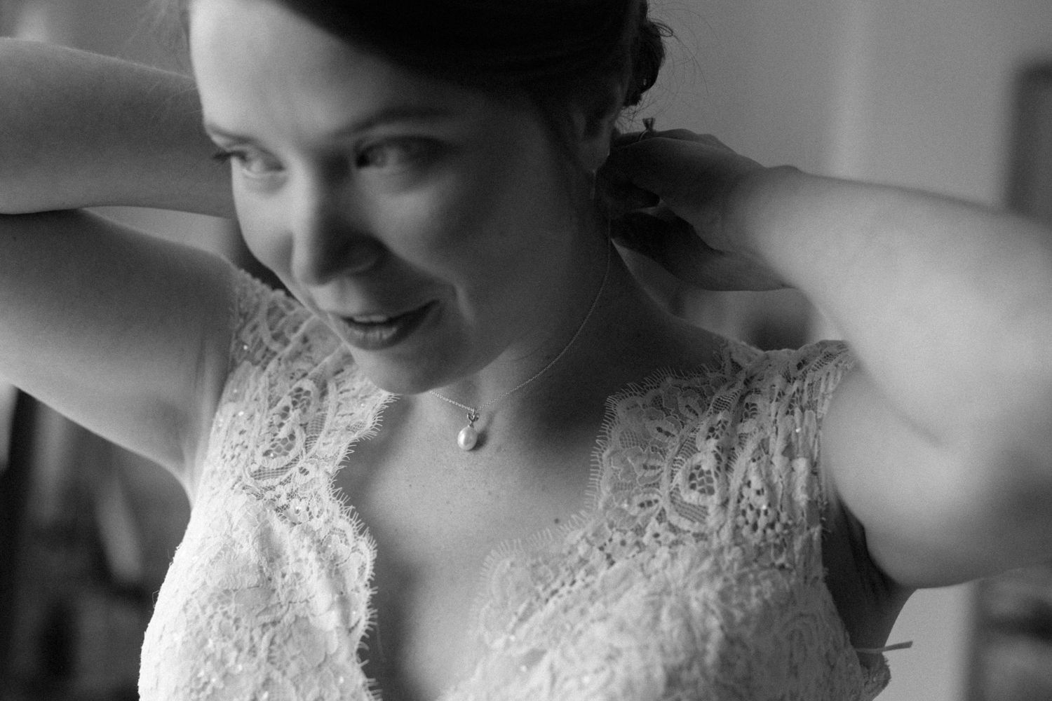 Black and white photo of a smiling bride in lace dress adjusting her necklace on her wedding day.
