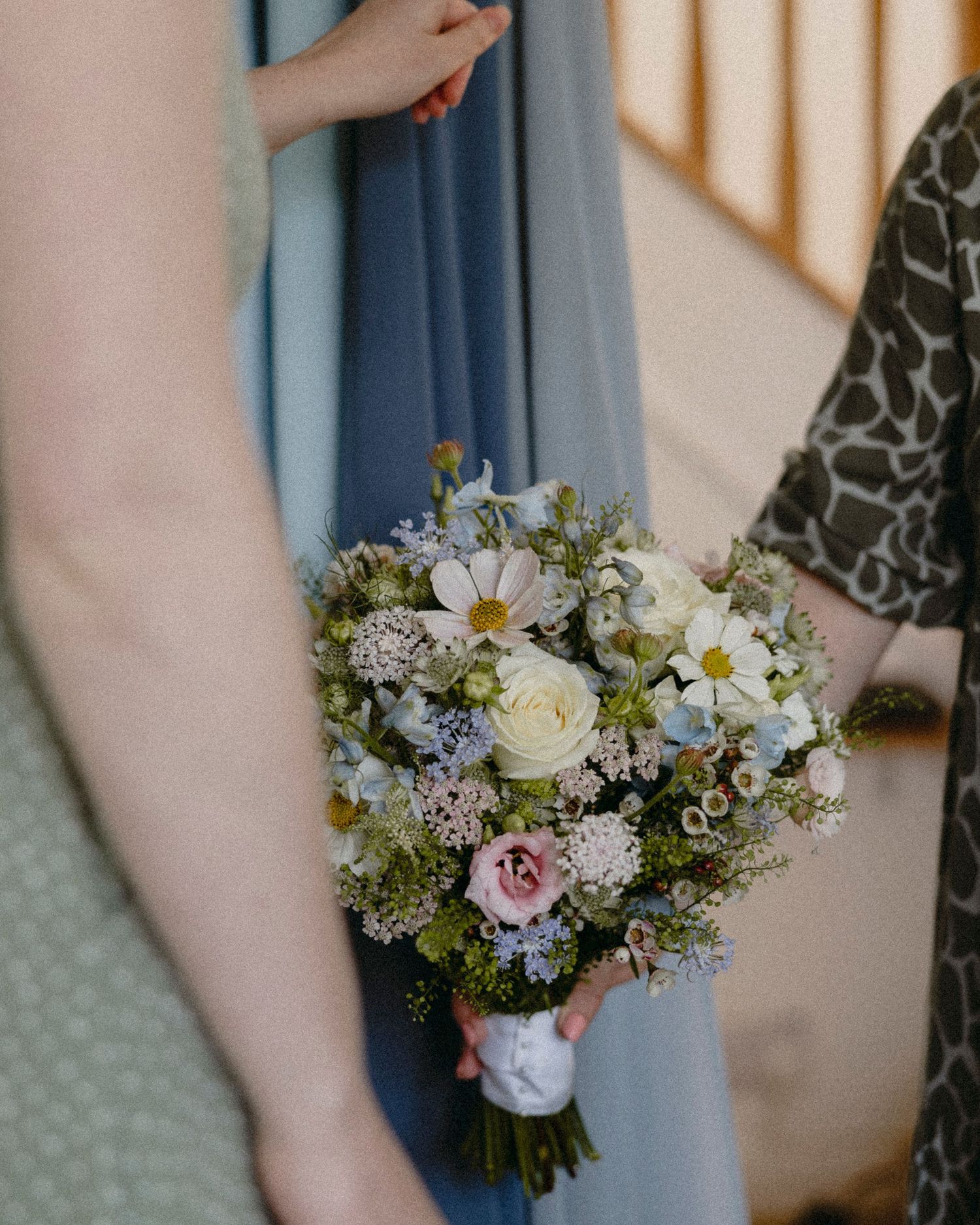 A bridesmaid holds a delicate wildflower bouquet with white, pink roses, daisies, and greenery at a wedding.