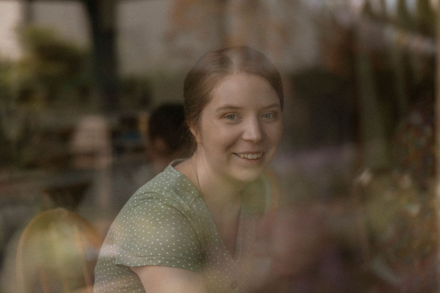 Smiling young woman in green dress seen through a window reflection in a cozy cafe setting.