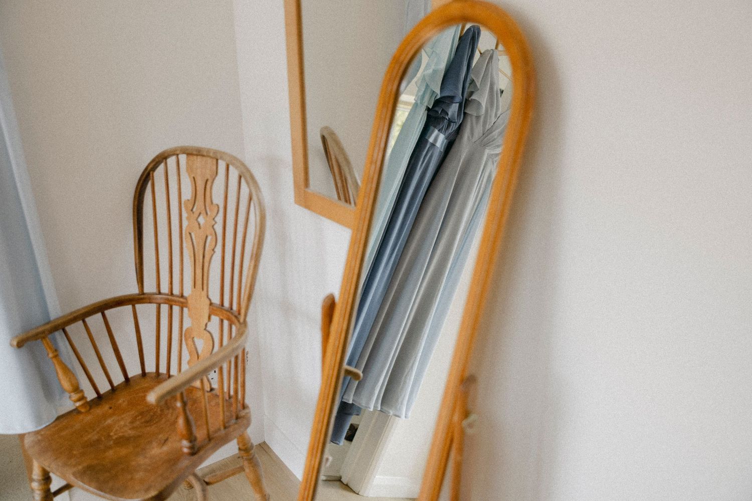 Wooden arched floor mirror leaning against white wall beside a Windsor chair in a minimalist room.