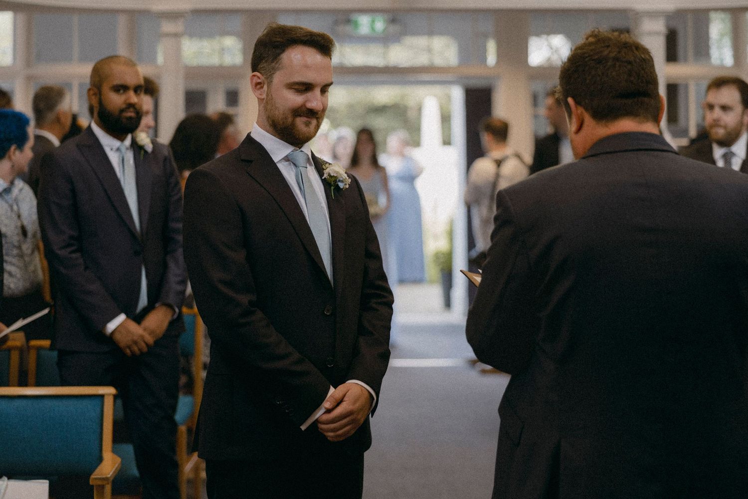 Groom in dark suit with gray tie smiling at officiant during indoor wedding ceremony with guests seated in background.