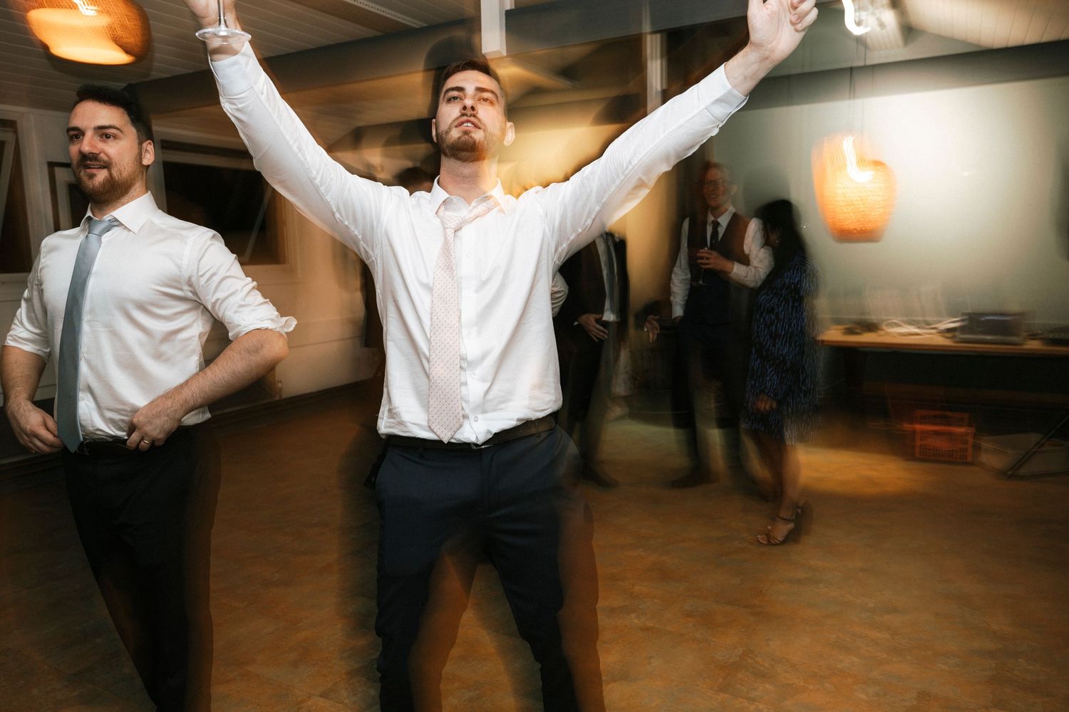 Men dancing energetically at a wedding reception, with one raising his arms in celebration on the dance floor.
