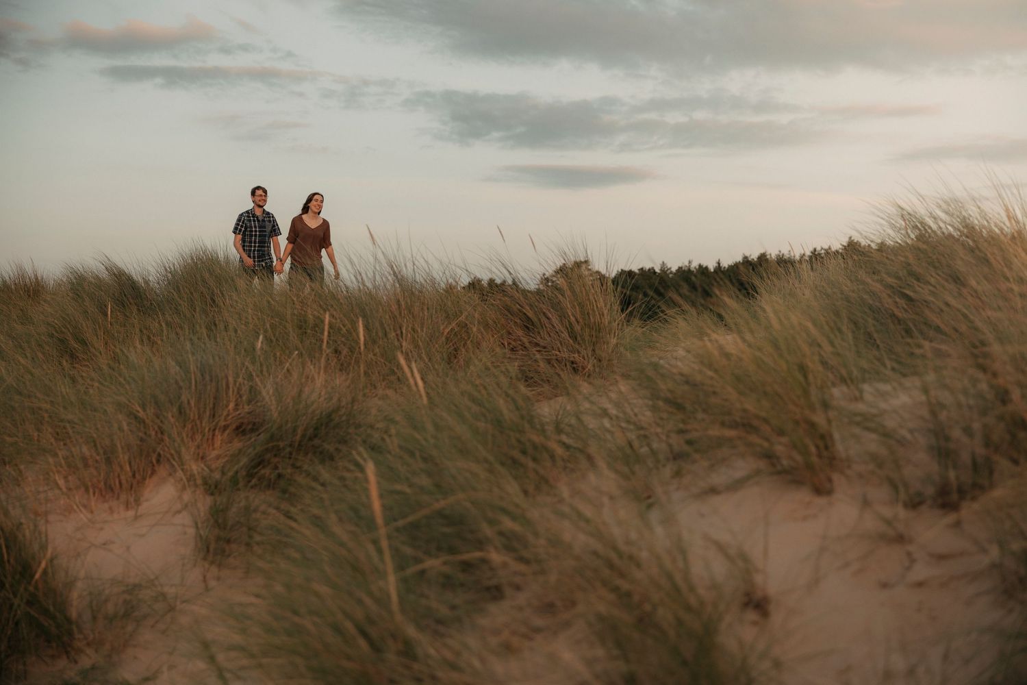 Couple standing on sandy dunes covered in tall beach grass under a cloudy evening sky.