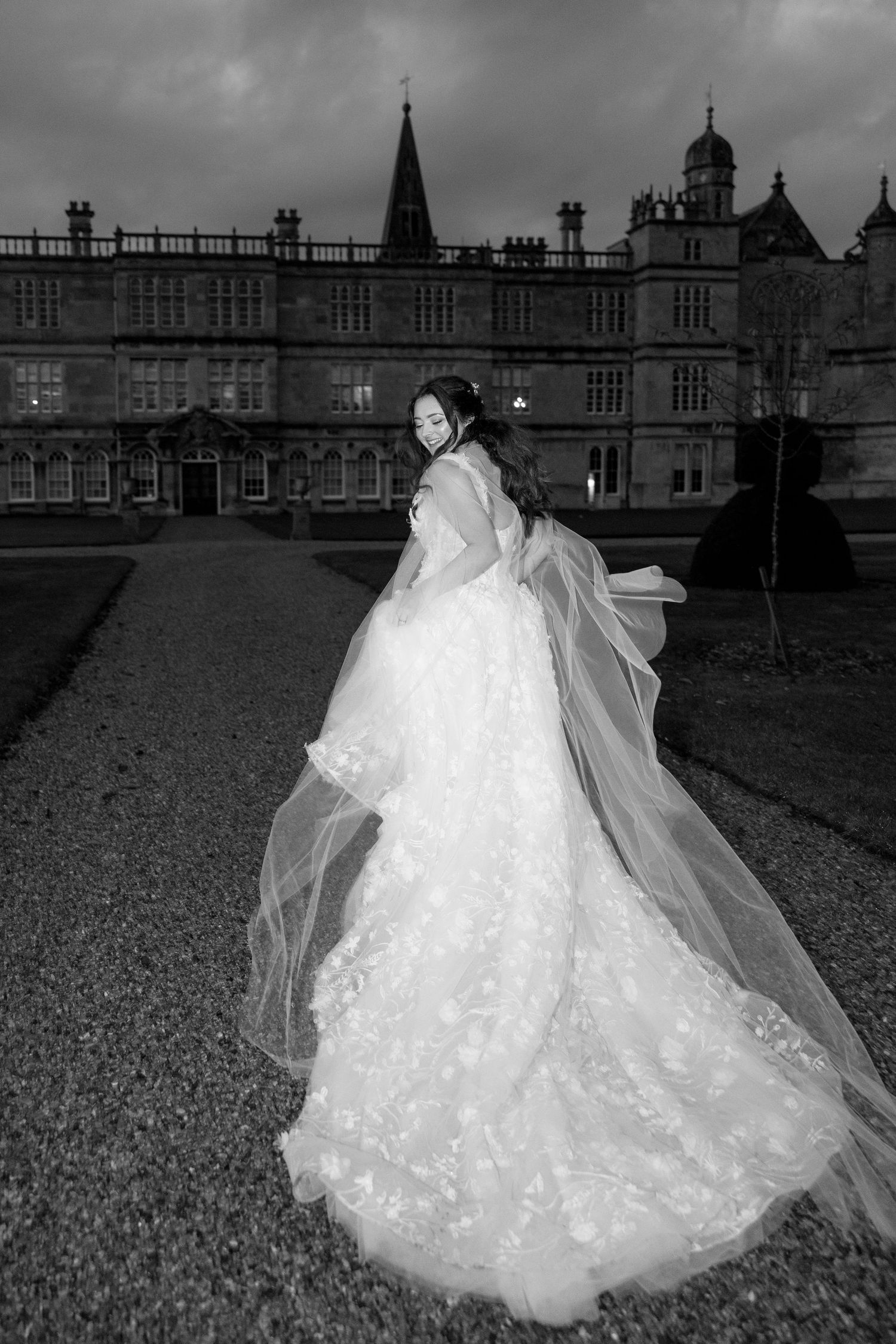 Black and white photo of a bride in a flowing lace gown with veil, posing before a grand historic manor.