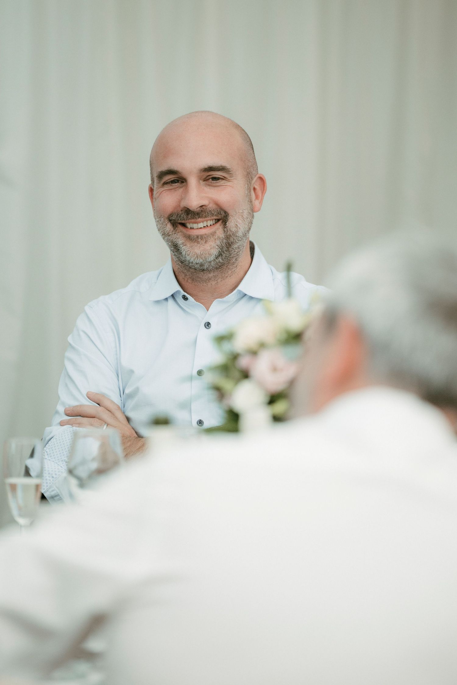Smiling bald man with beard in light blue shirt seated at elegant dining table with floral centerpiece.