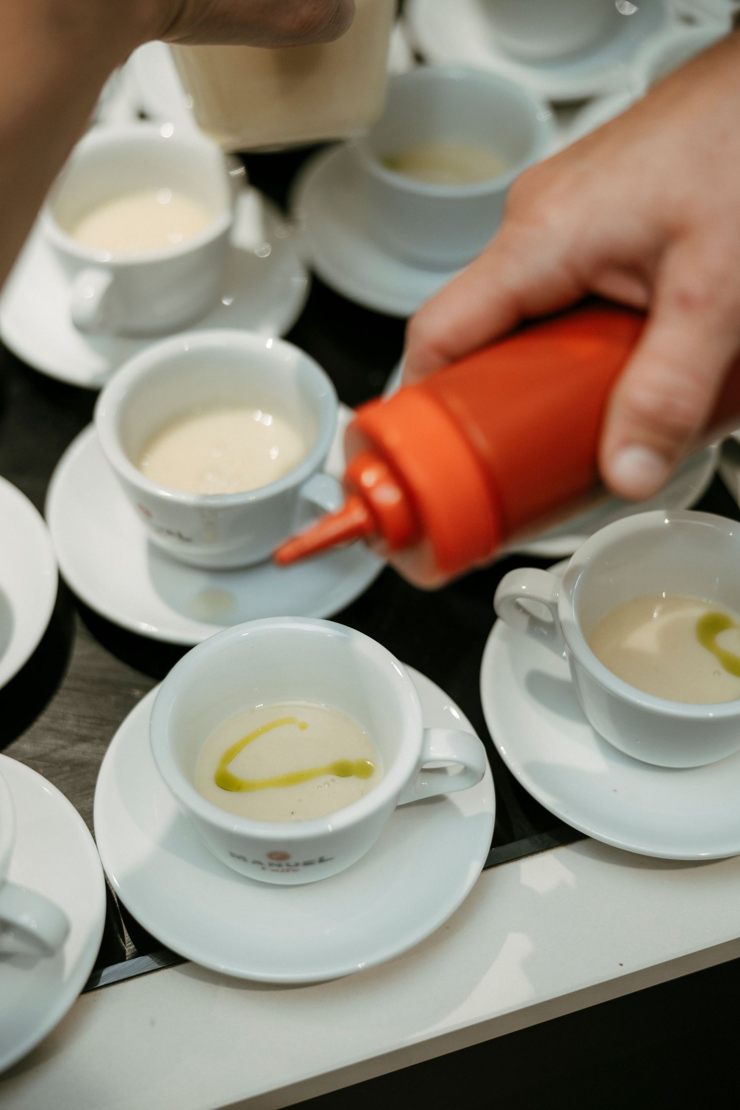Hand pouring olive oil from orange squeeze bottle into white espresso cups on saucers arranged on a table.