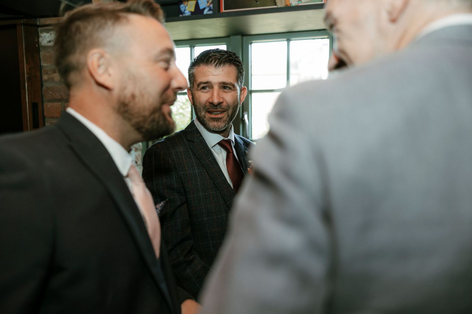 Three men in suits smiling and conversing at a formal event or wedding reception indoors.