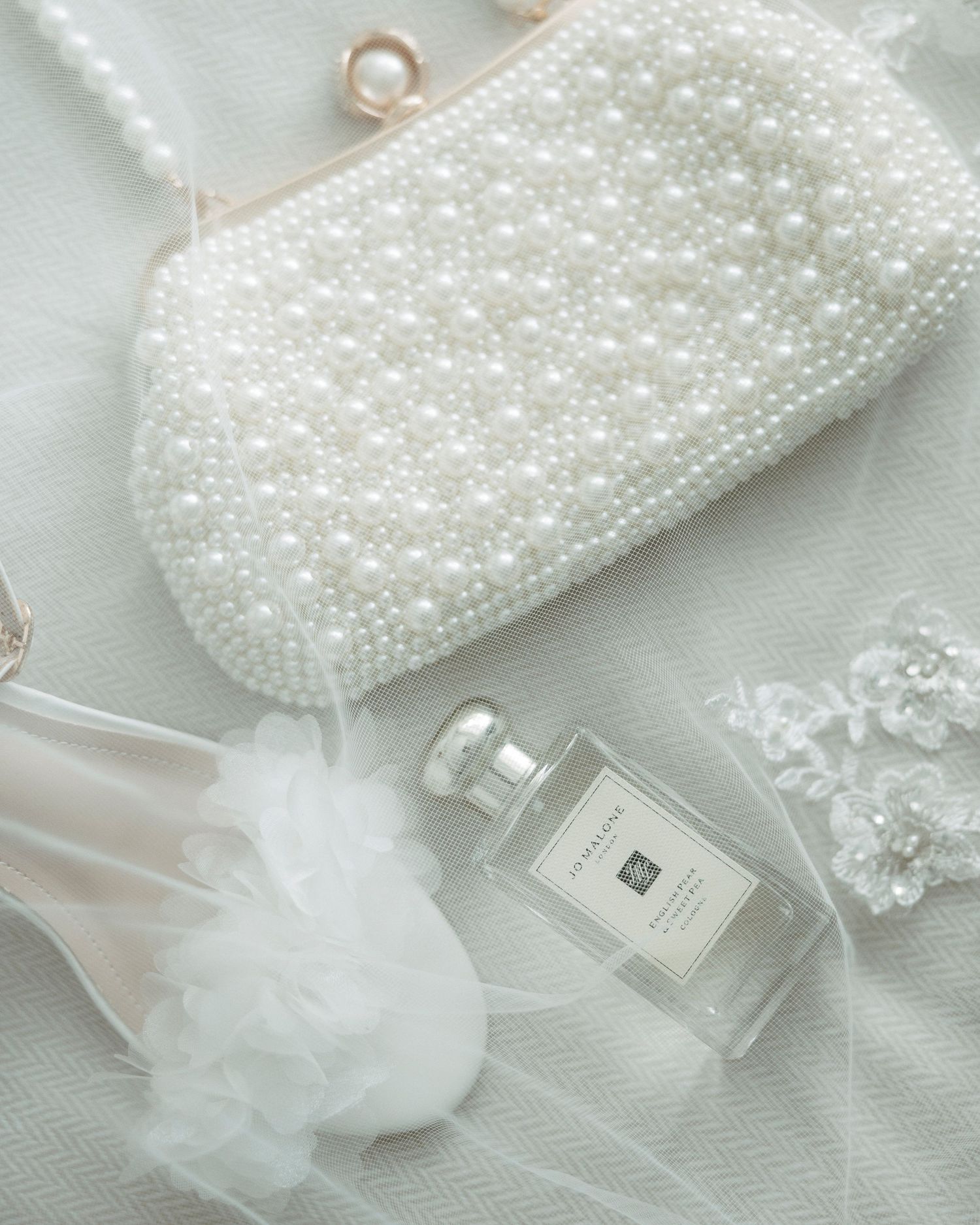 Pearl-beaded bridal clutch bag with Jo Malone perfume, floral hair accessories, and white tulle on ivory fabric.