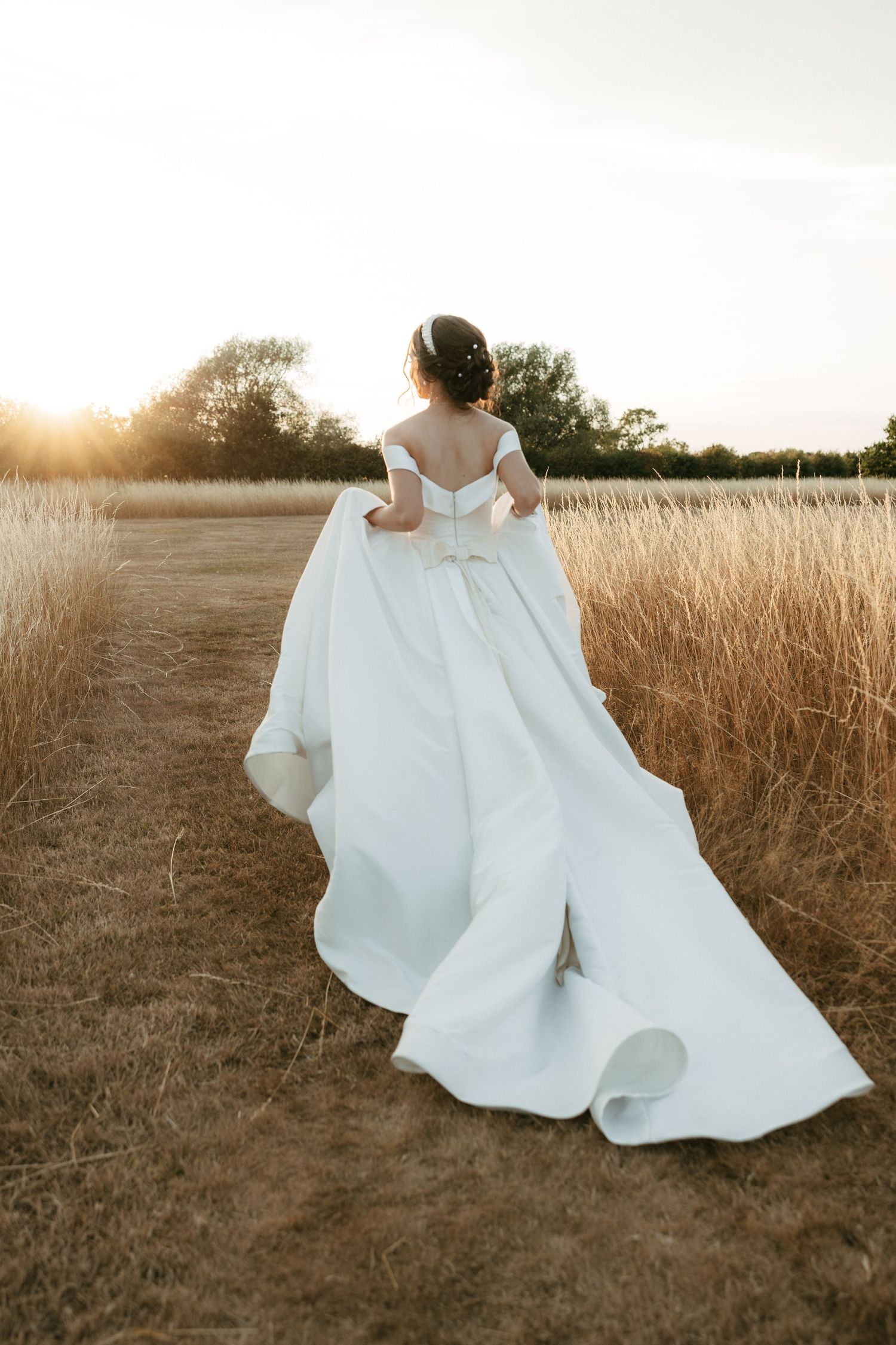 Bride in elegant off-shoulder white gown with long train walks through golden field at sunset.
