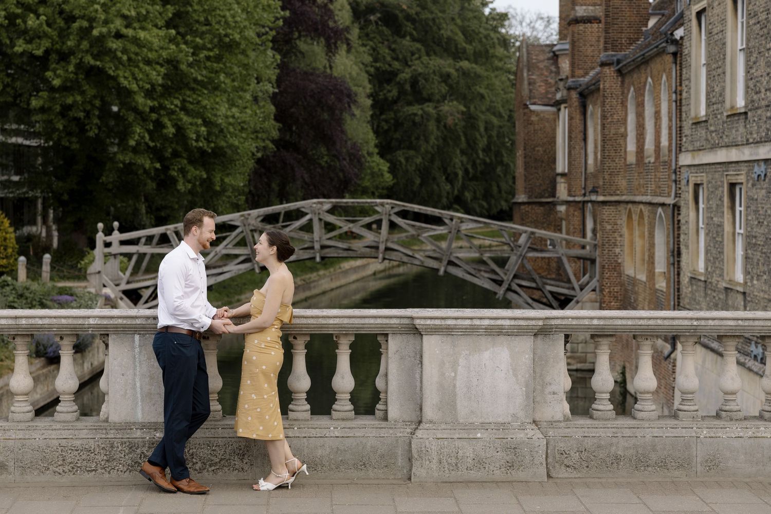 Couple holding hands by Mathematical Bridge in Cambridge, England, surrounded by historic architecture.