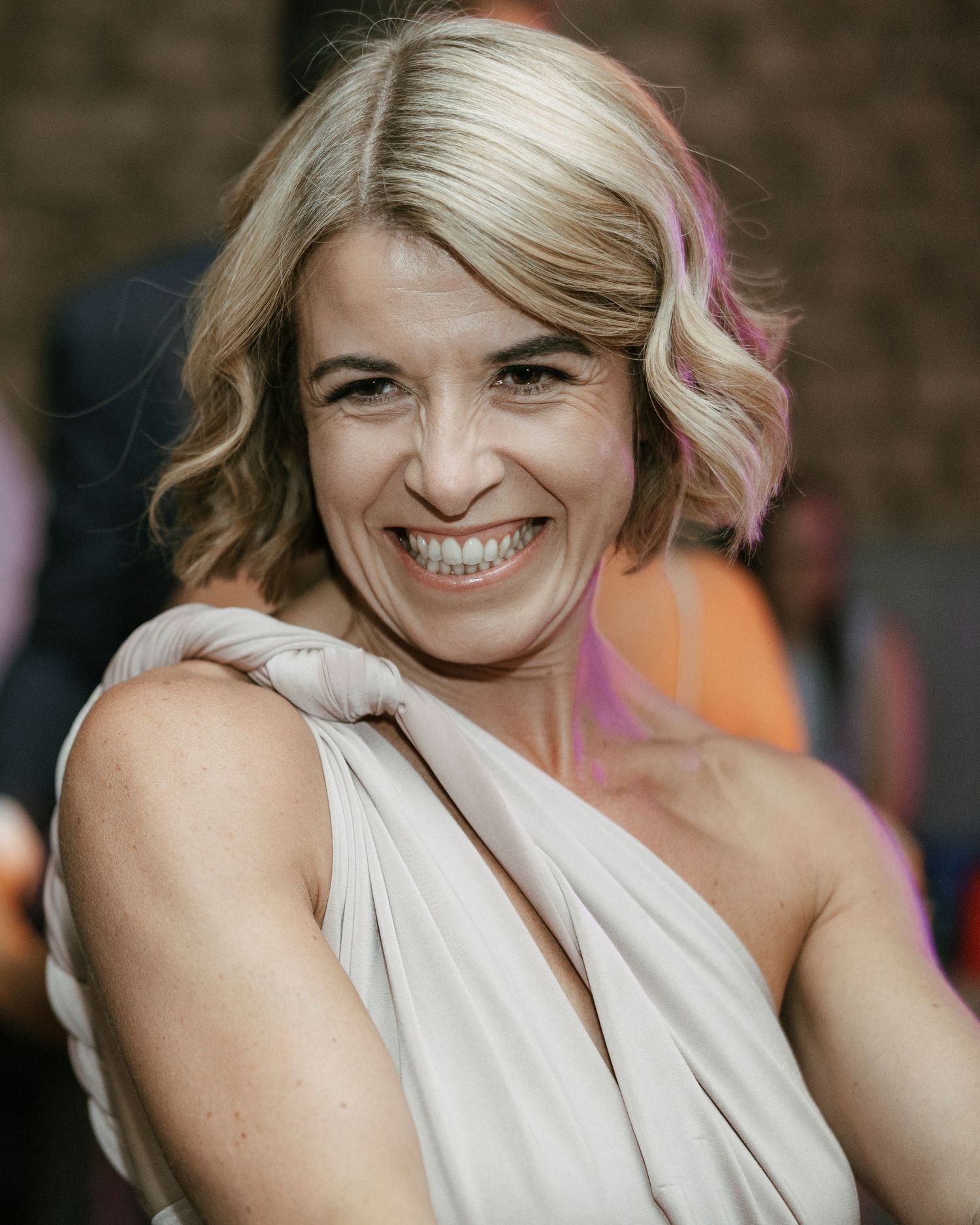Smiling blonde woman in elegant white draped dress at wedding celebration.