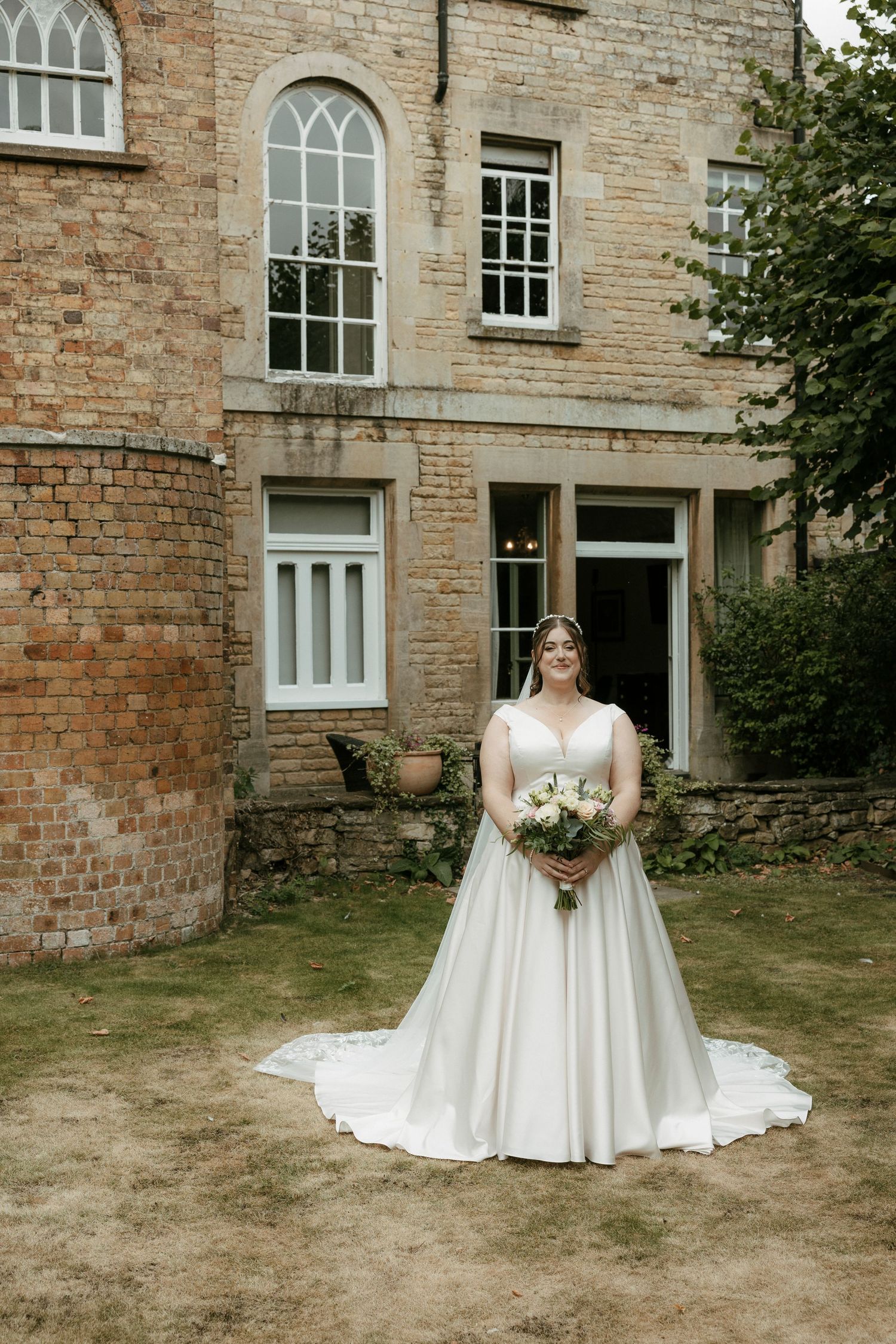 Bride in satin ball gown holding white bouquet outside historic brick manor.