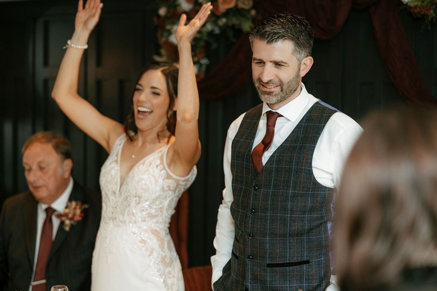 A joyful couple celebrates during their wedding reception, with the bride raising her arms and the groom smiling beside her.