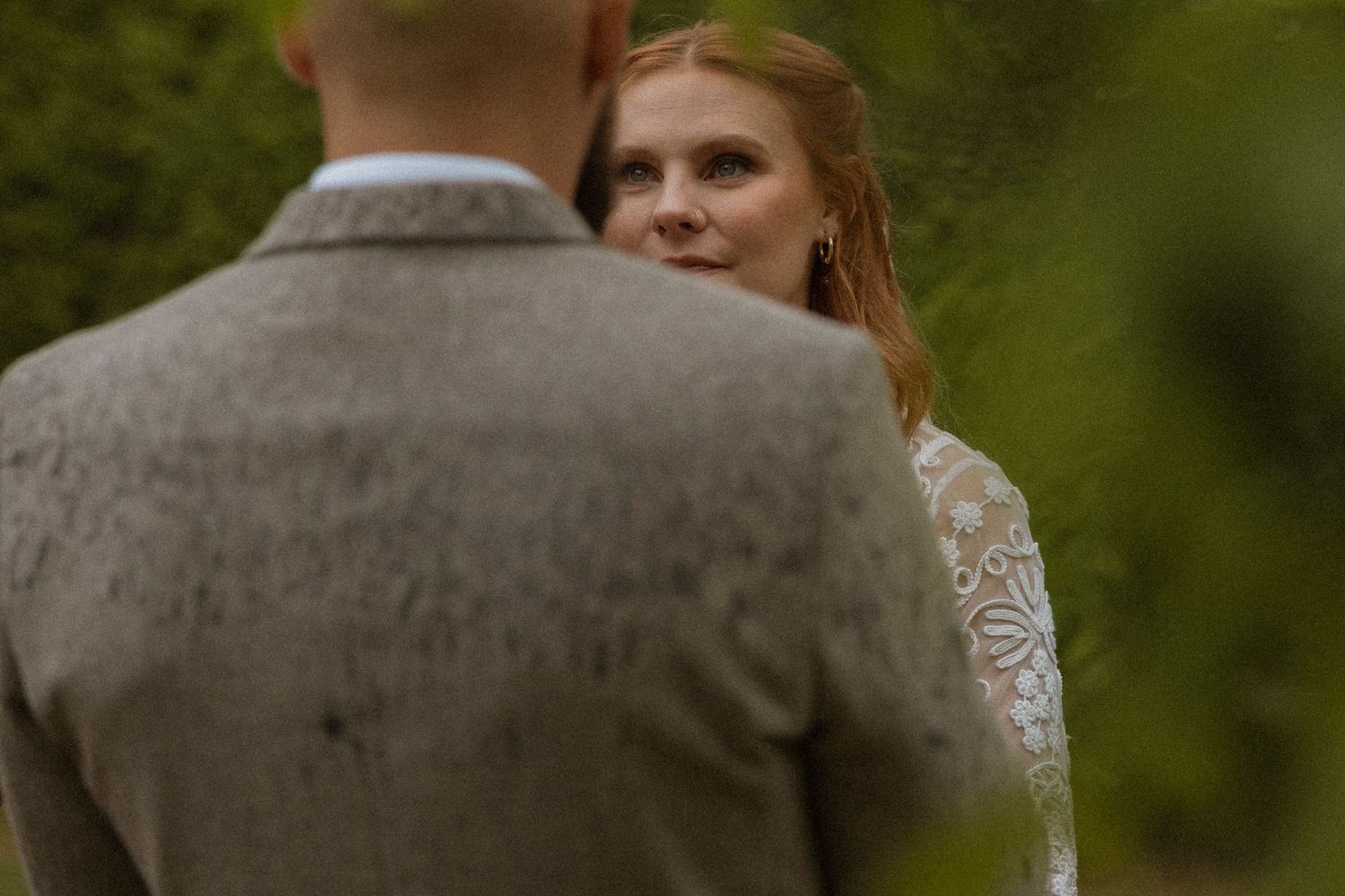 Bride in lace wedding dress gazes lovingly at groom in gray suit outdoors during wedding ceremony.
