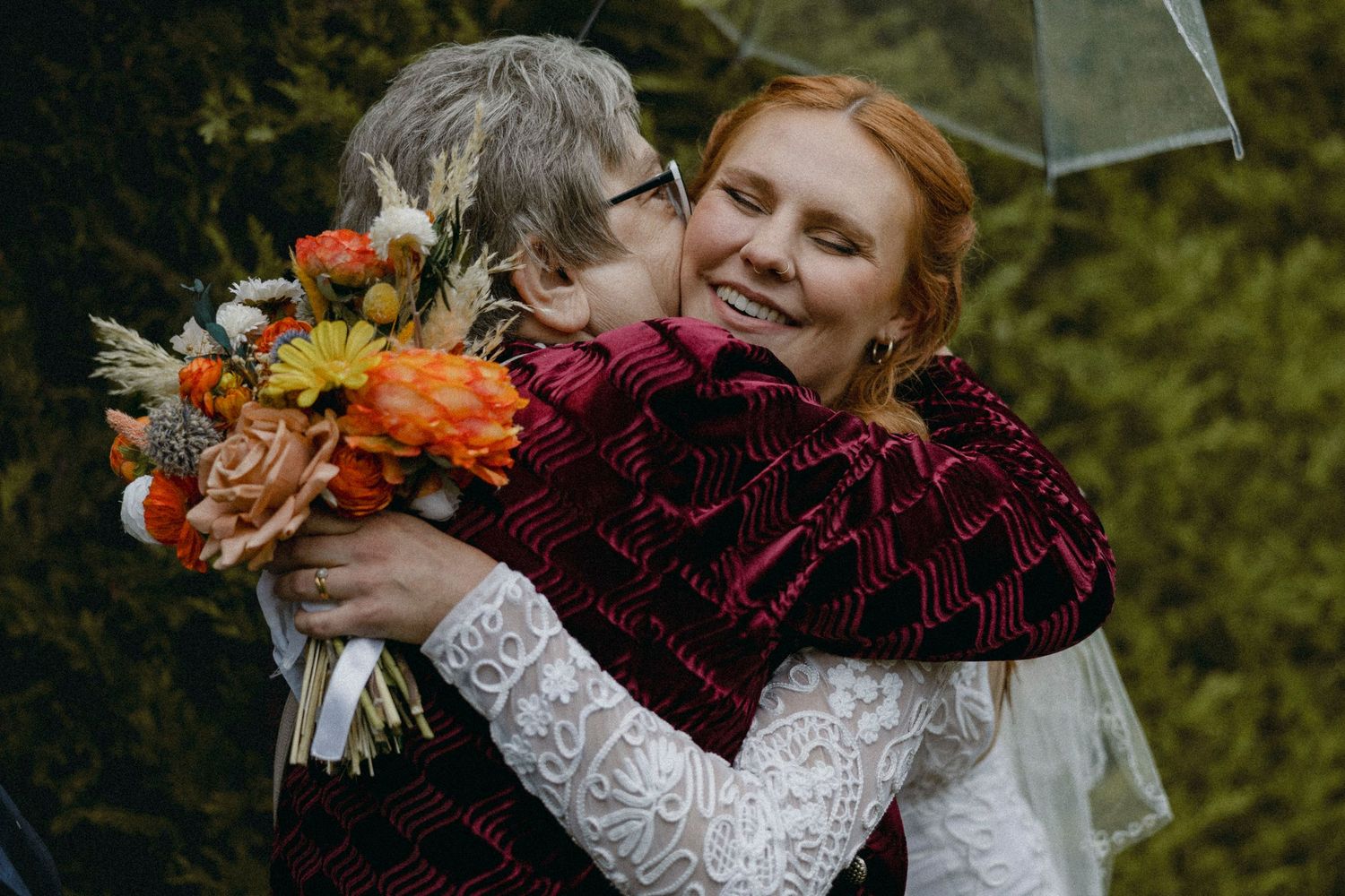 Bride in white lace dress hugging older woman in red cardigan, holding orange autumn wedding bouquet outdoors.