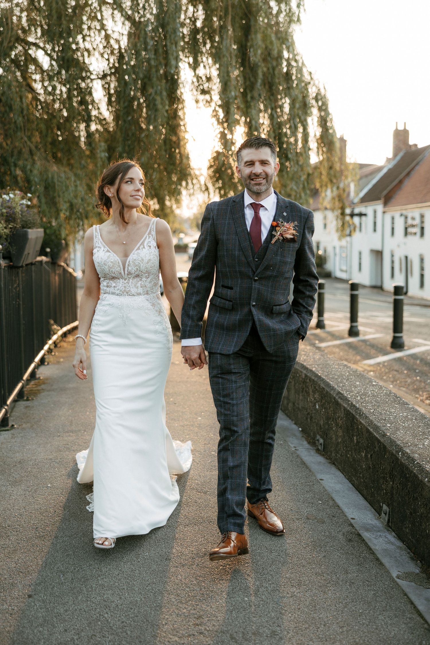 Bride and groom walking hand in hand on a cobblestone street at golden hour, wearing elegant wedding attire.