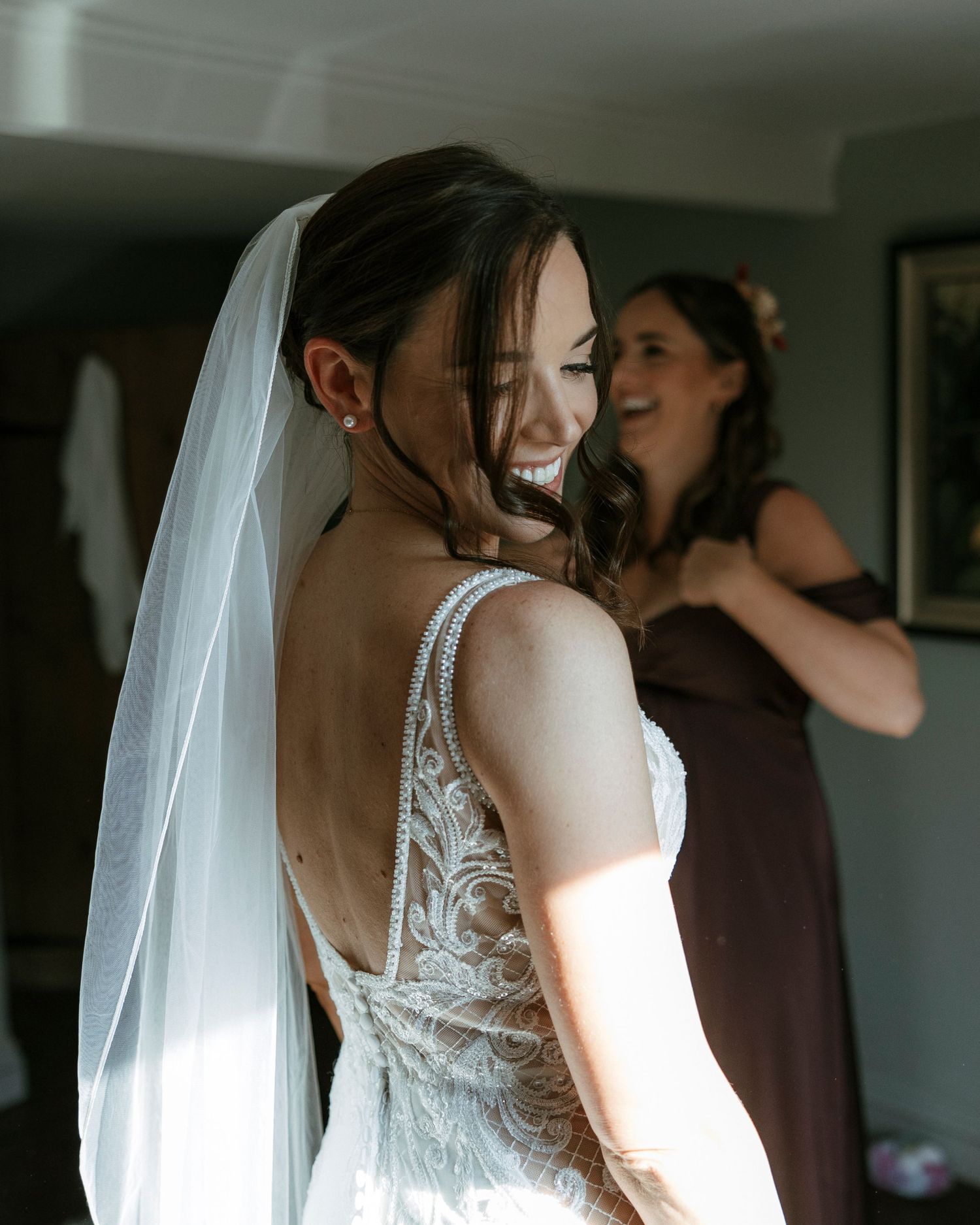Bride in lace backless wedding gown and veil smiles over shoulder while bridesmaid in purple dress assists her.