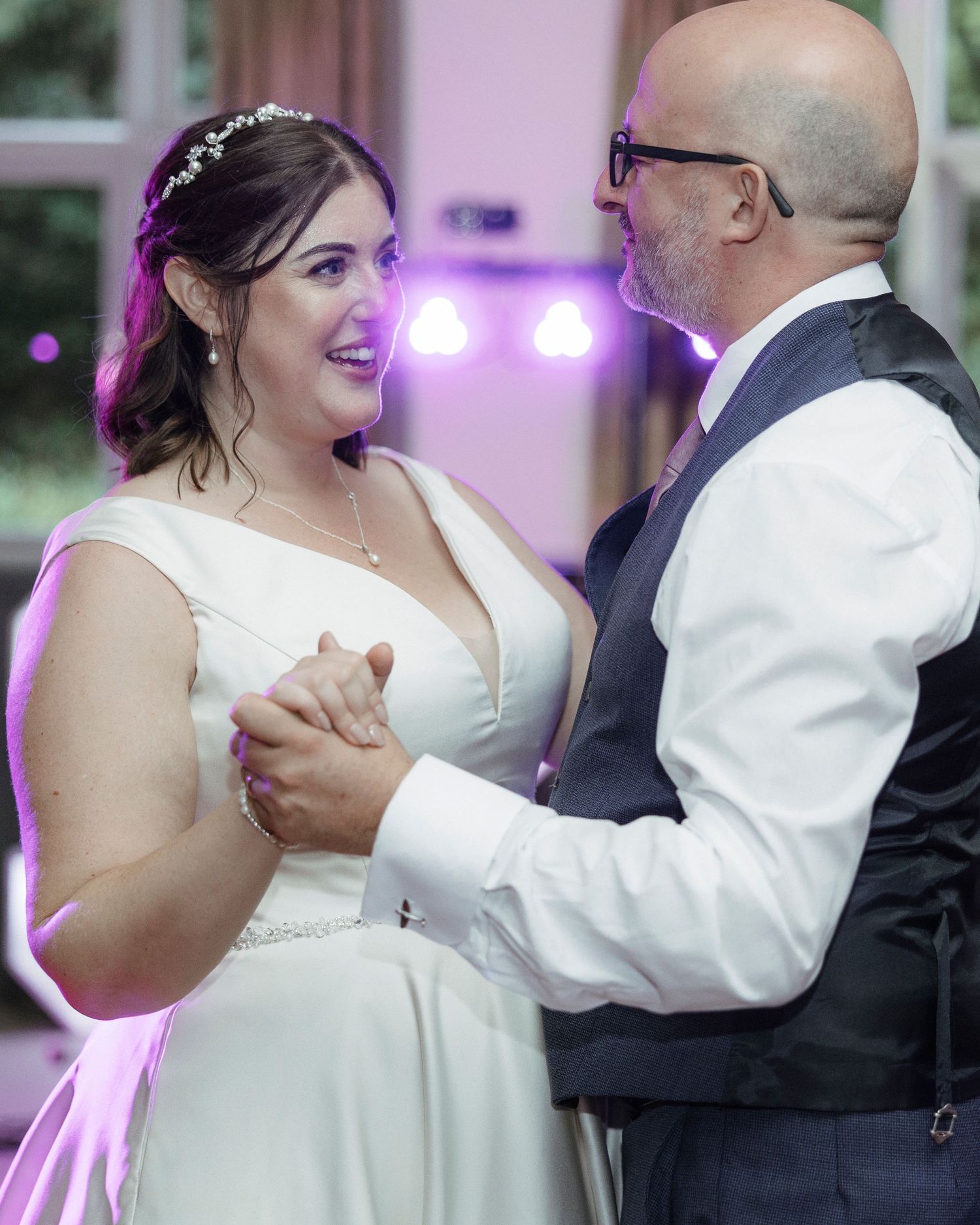 Bride and groom share an intimate first dance, smiling at each other under purple reception lights.