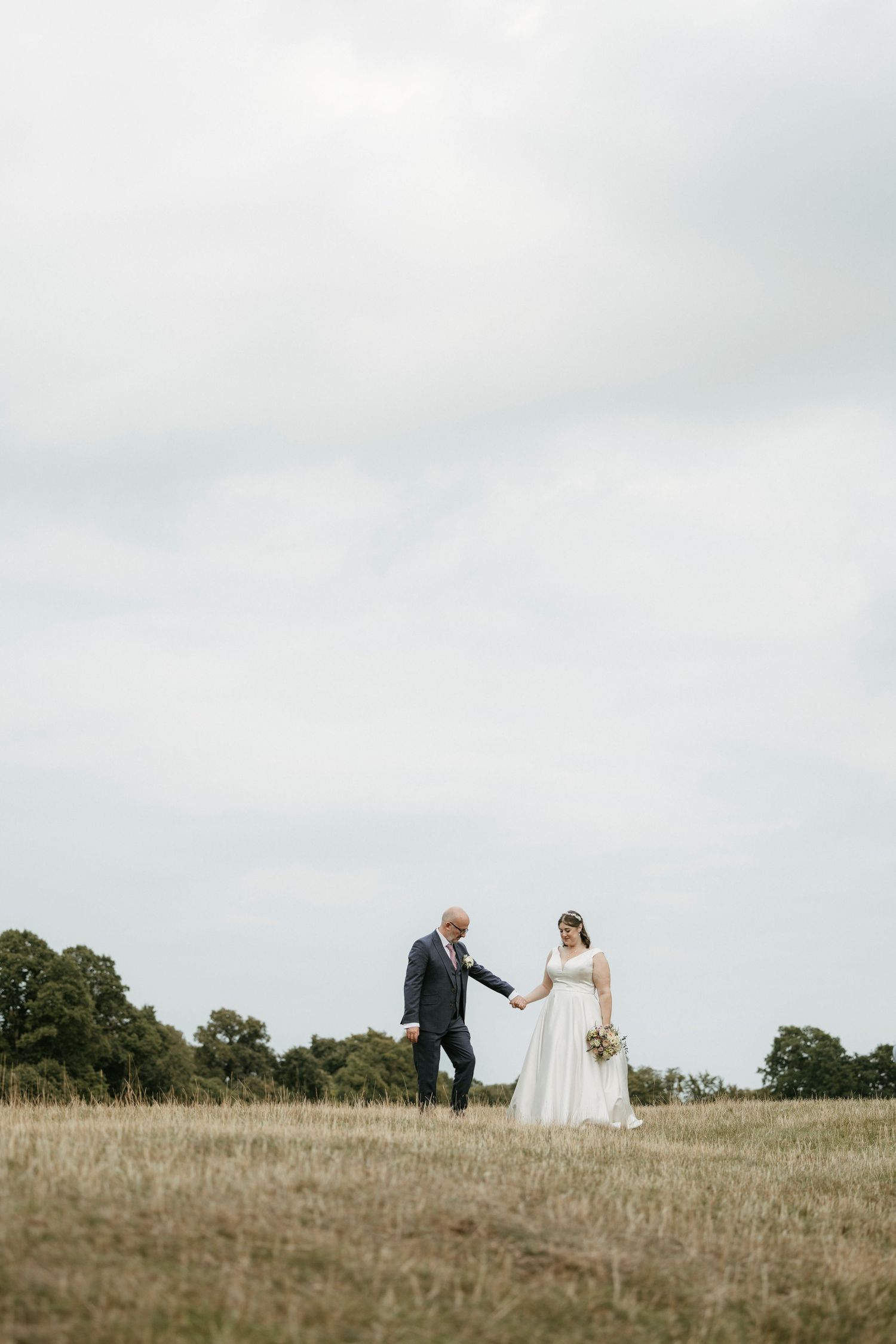 Bride and groom holding hands walking through golden field on cloudy day in elegant wedding attire.