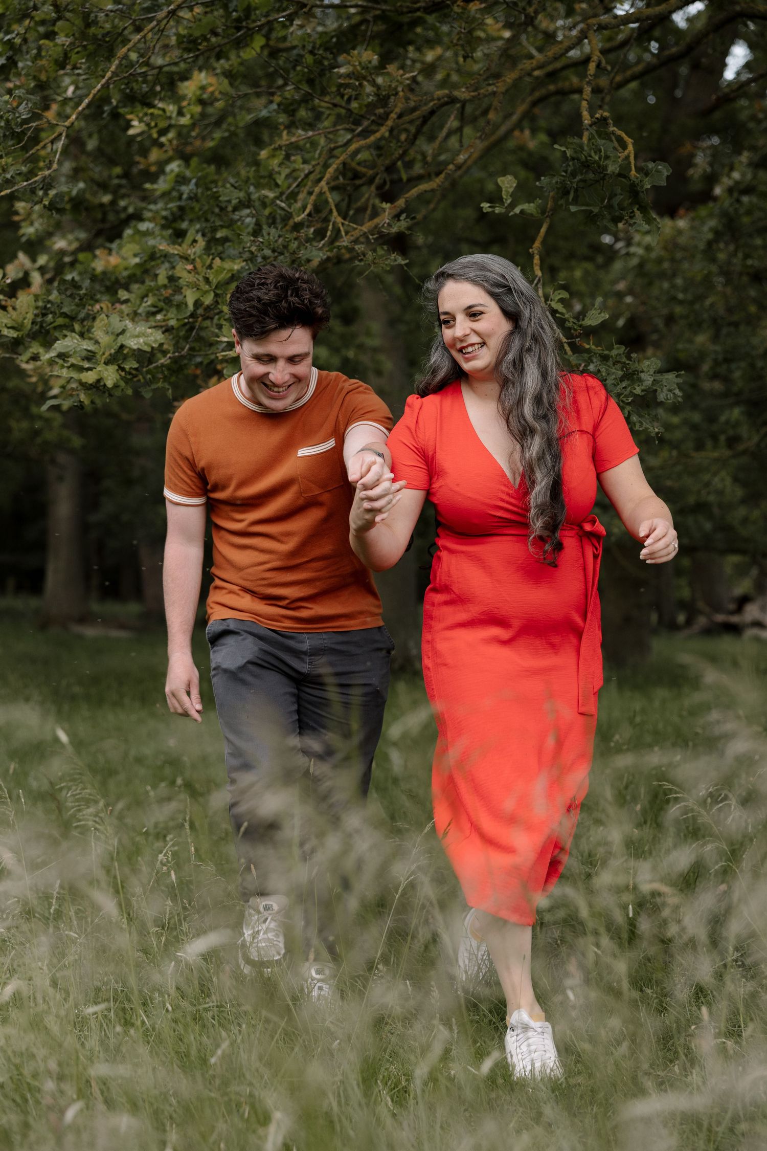 A smiling couple holds hands while walking through a lush green field, wearing red and orange outfits.