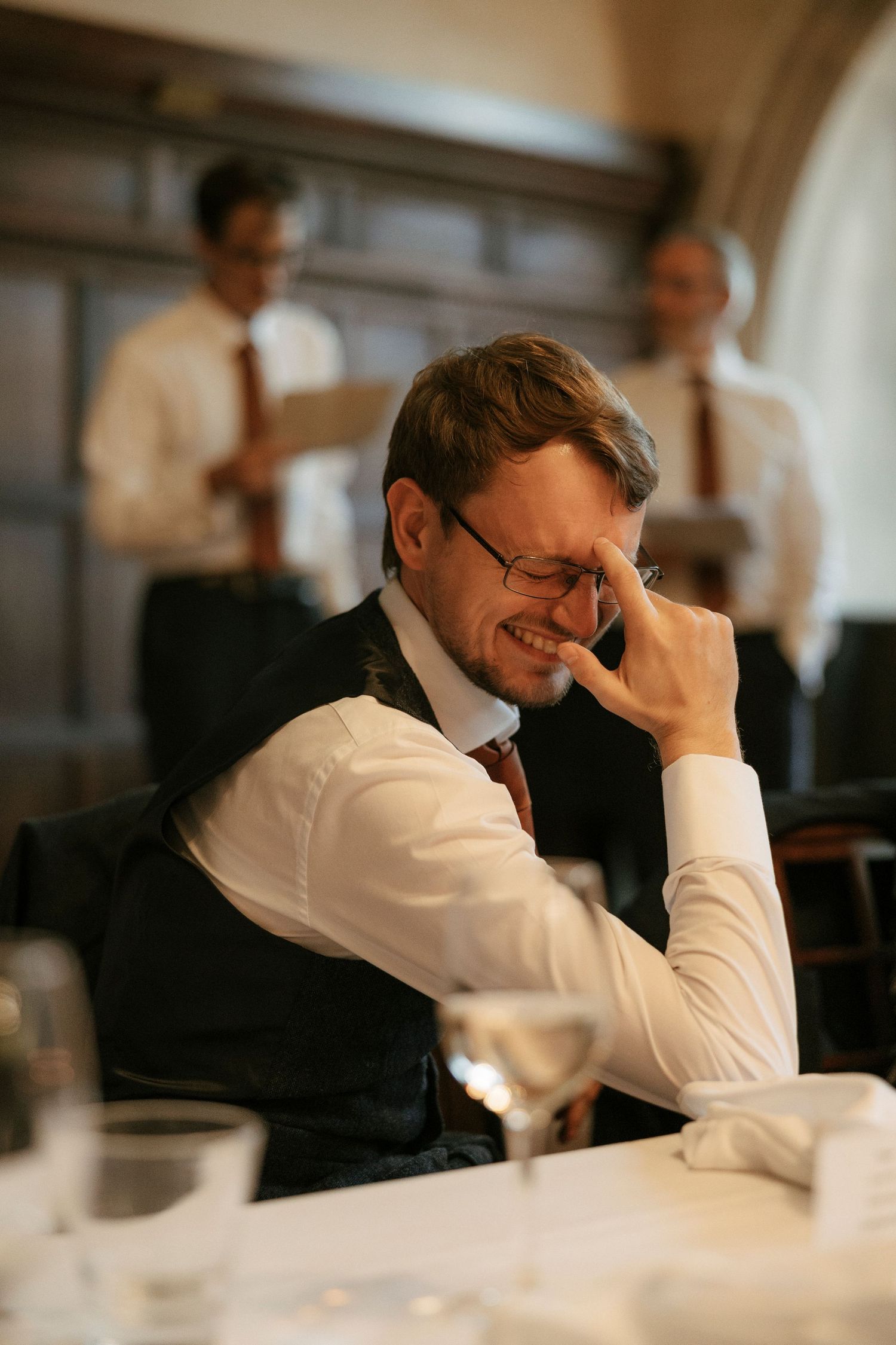 Man in vest and glasses laughing at a table during a formal dinner event with colleagues in background.