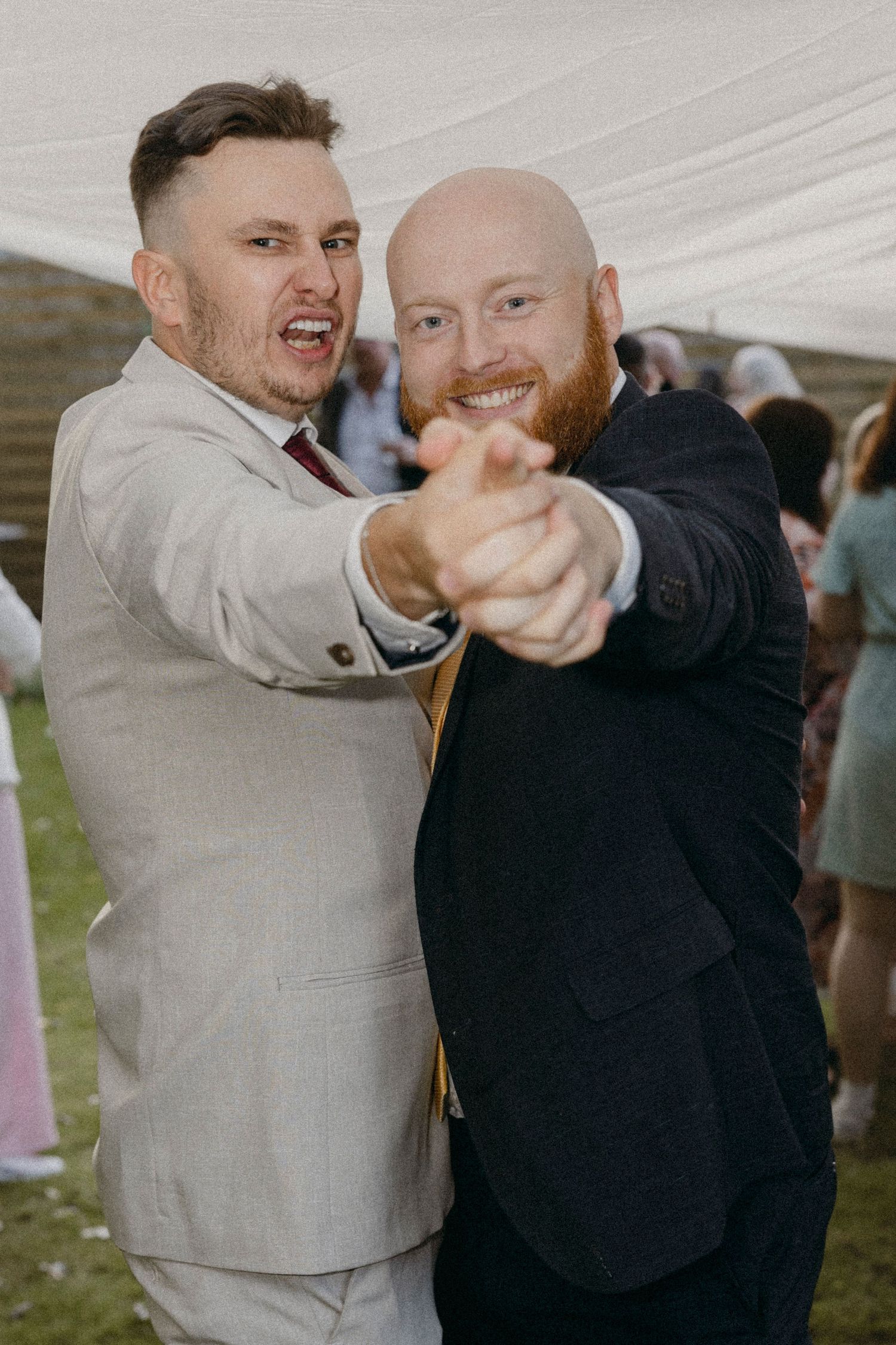 Two men in suits smiling and pointing at camera at a wedding reception under a marquee tent.
