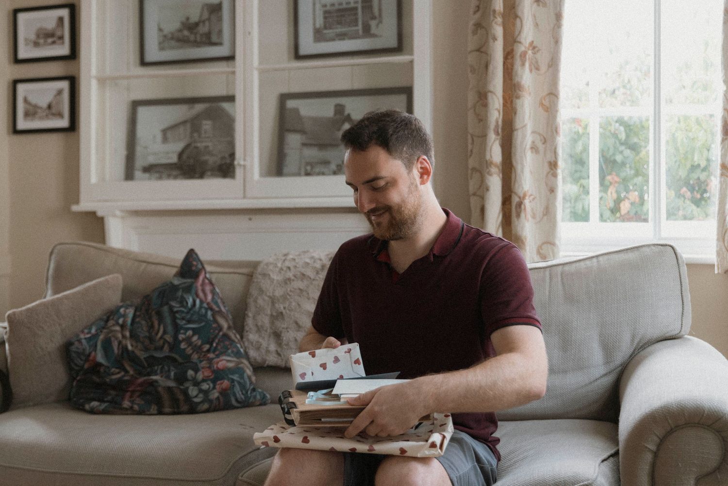 Man sitting on a sofa looking through old photos in a cozy living room with framed pictures on the wall.