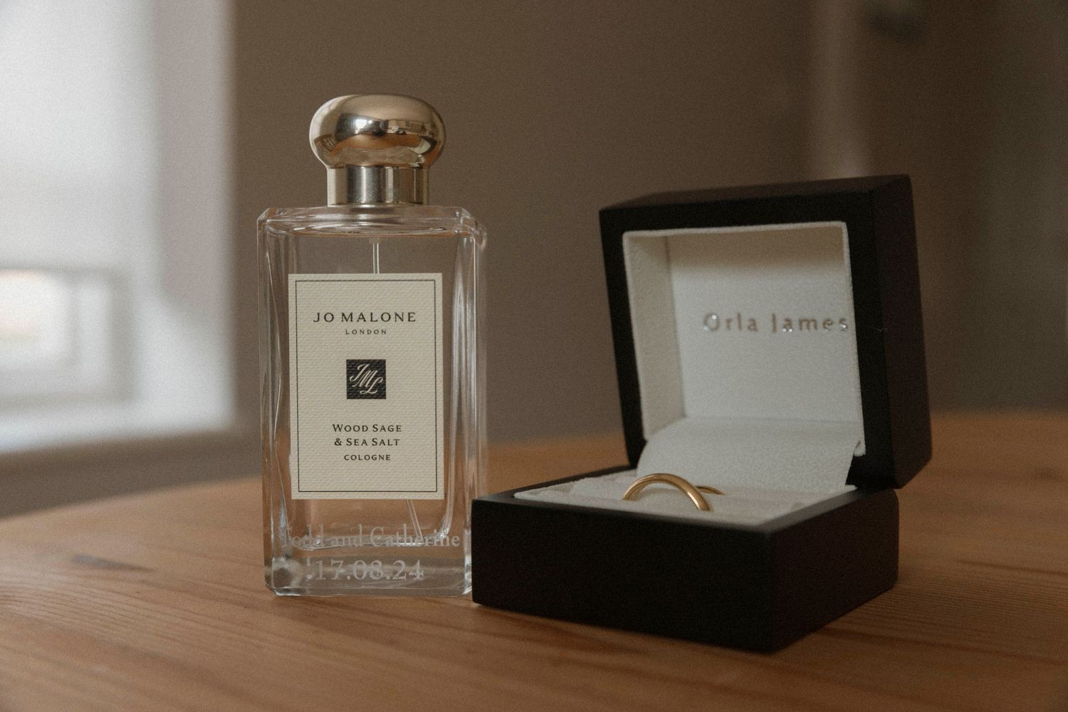 Jo Malone London perfume bottle beside a gold wedding ring in a black Gigi Jewels ring box on a wooden table.