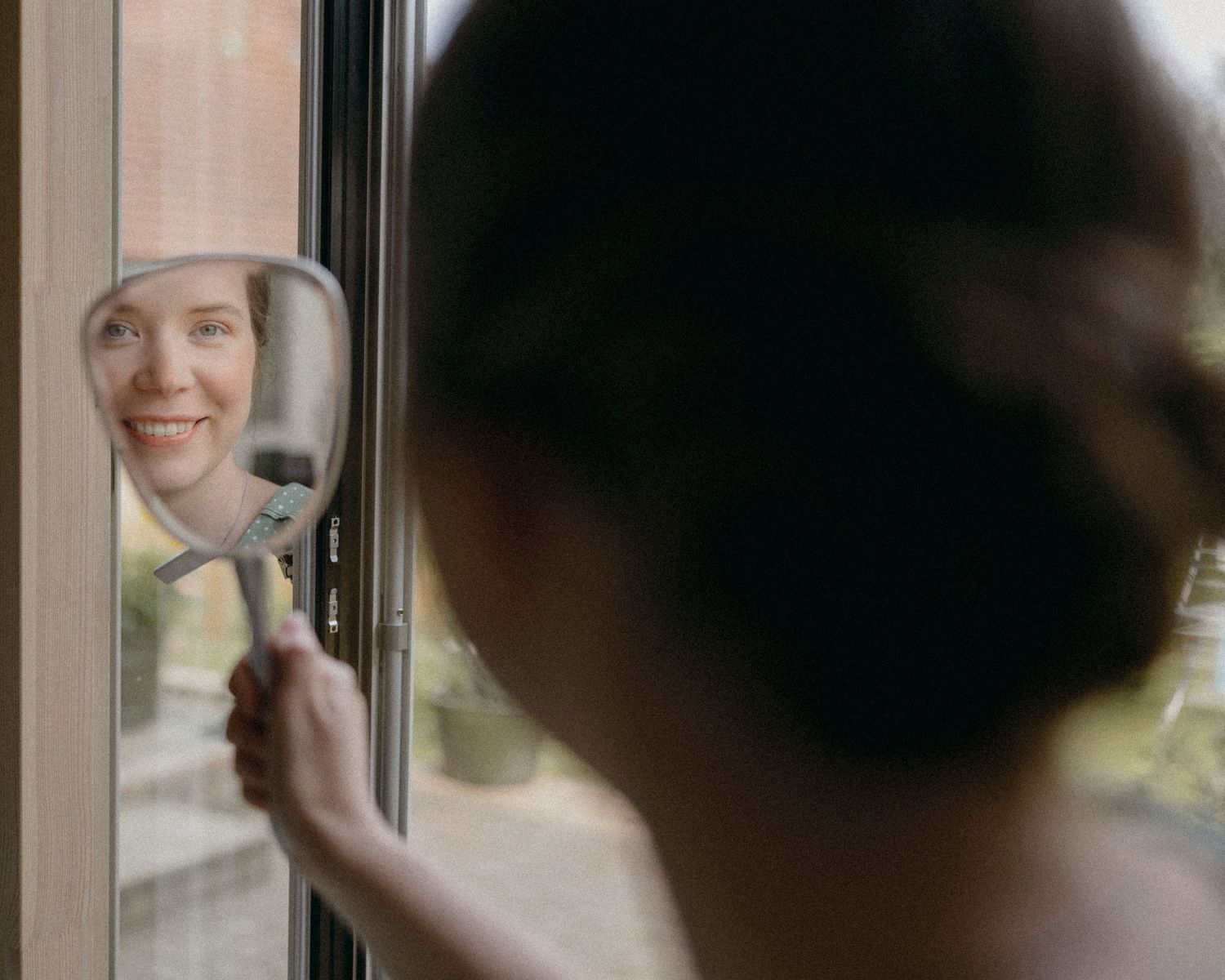 A smiling woman views her reflection in a handheld mirror held up near a window.